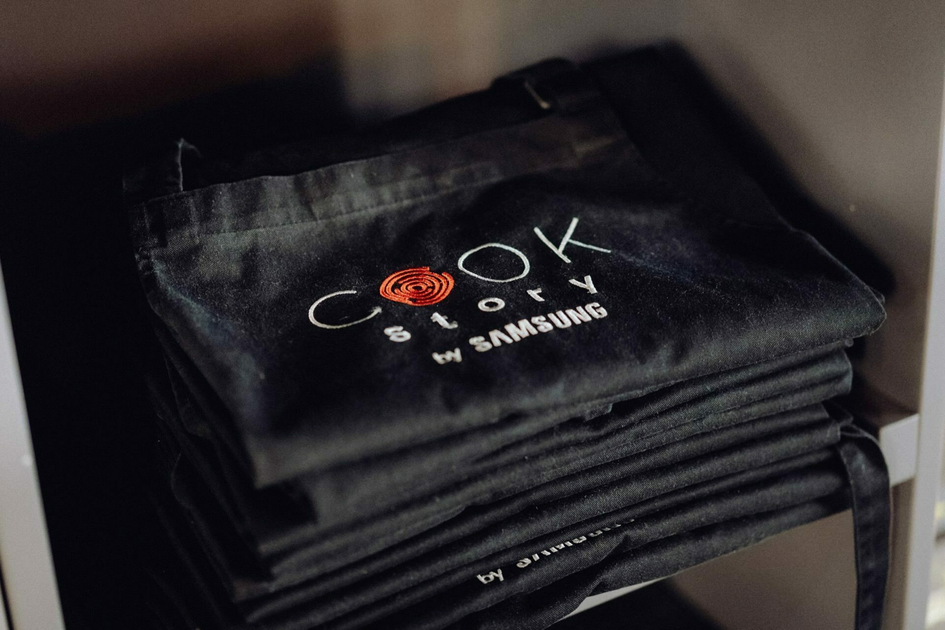 On the shelf is a neatly stacked stack of black aprons embroidered with the "Cook Story by Samsung" logo. The logo, made by an expert event photographer Warsaw, features the word "Cook" with an orange swirl integrated with the letter "O" and the words "Story by Samsung" below it. 