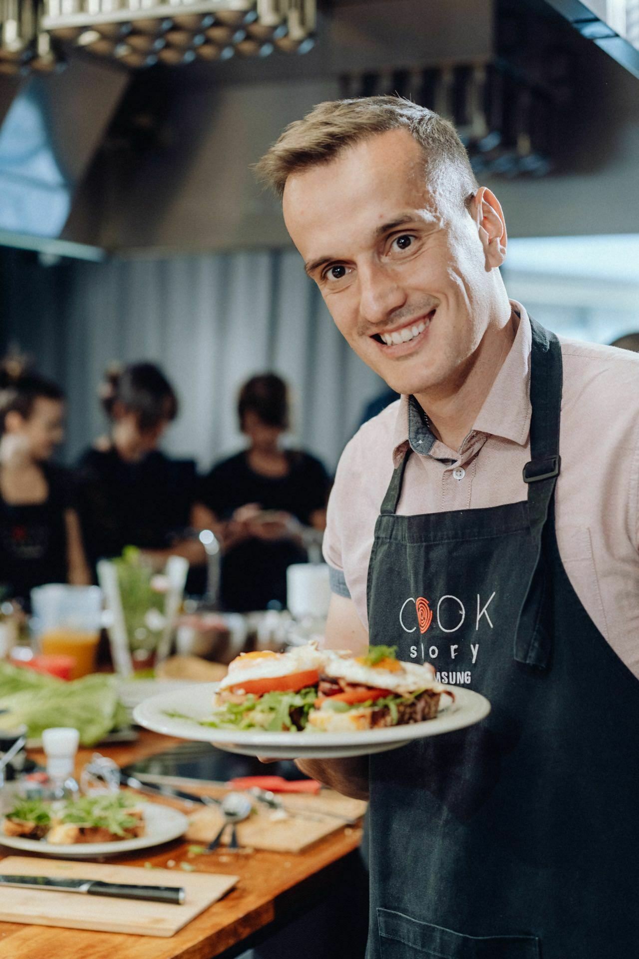 A smiling person dressed in an apron holds a plate with a sandwich, standing in a kitchen with a wooden countertop and various utensils. Blurred figures can be seen in the background, suggesting a photo essay of an event or a cooking course. 