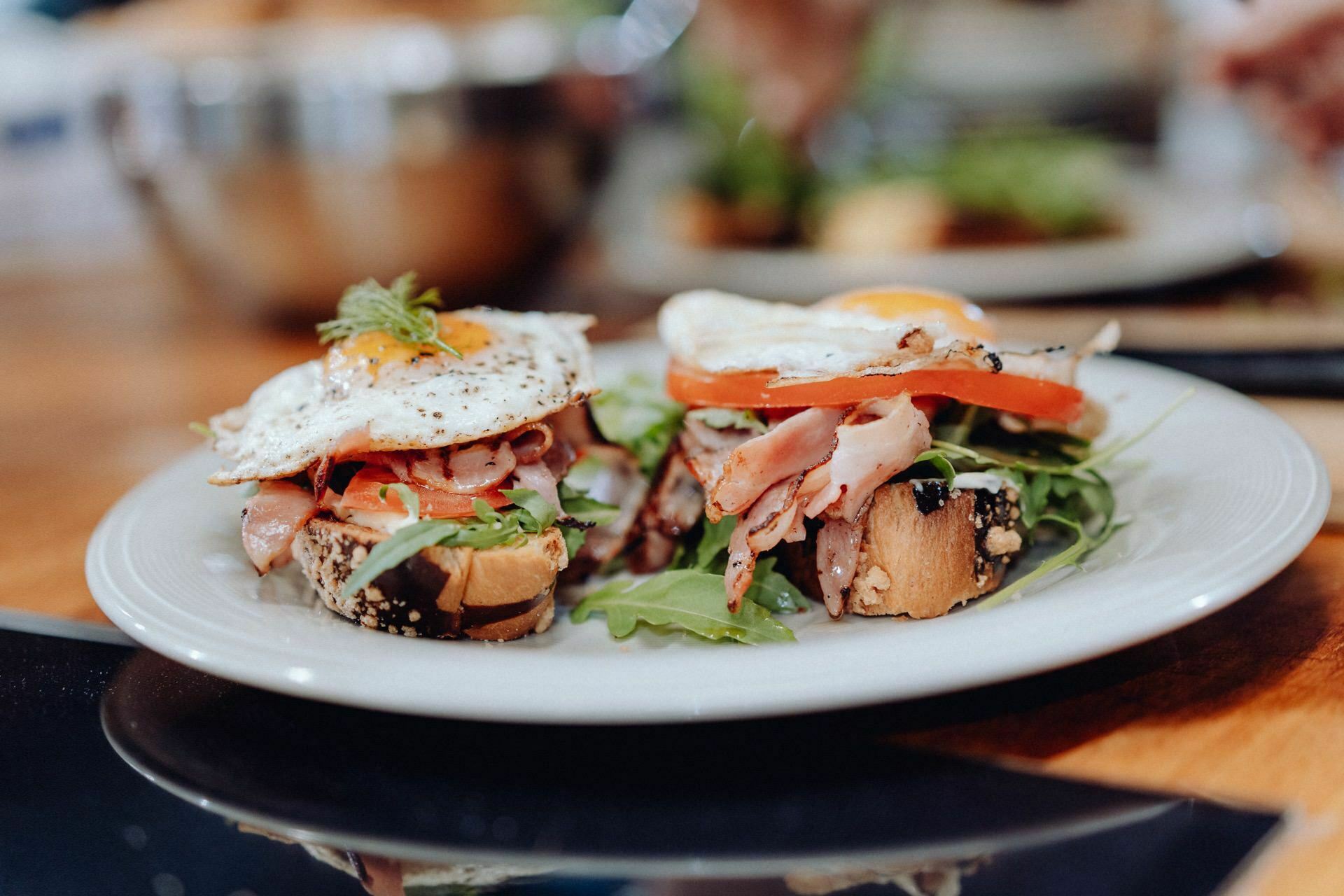 A plate with two servings of open-faced sandwiches topped with fried eggs, slices of ham, tomato and arugula, on crusty toasted seeded bread. In the background, you can see a blurry kitchen setup that could have come straight from an event photographer's photo shoot in Warsaw. 