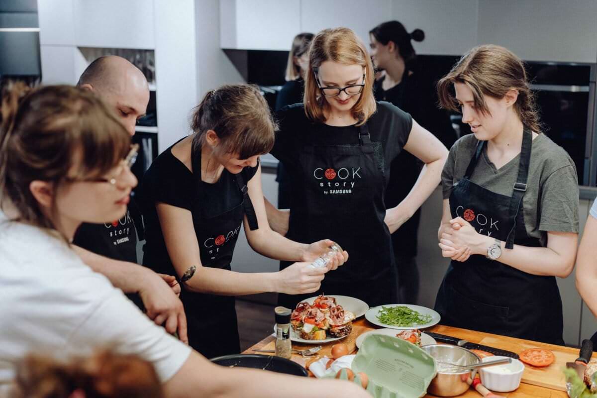 Grupa osób ubranych w czarne fartuchy zebrała się wokół blatu kuchennego, zajmując się gotowaniem. Jedna osoba wylewa na talerz śmietanę lub sos, inne zaś przyglądają się i biorą udział w przygotowywaniu różnych składników. Scena uchwycona w ramach fotografii eventowej sugeruje, że są to ożywione zajęcia lub warsztaty kulinarne.