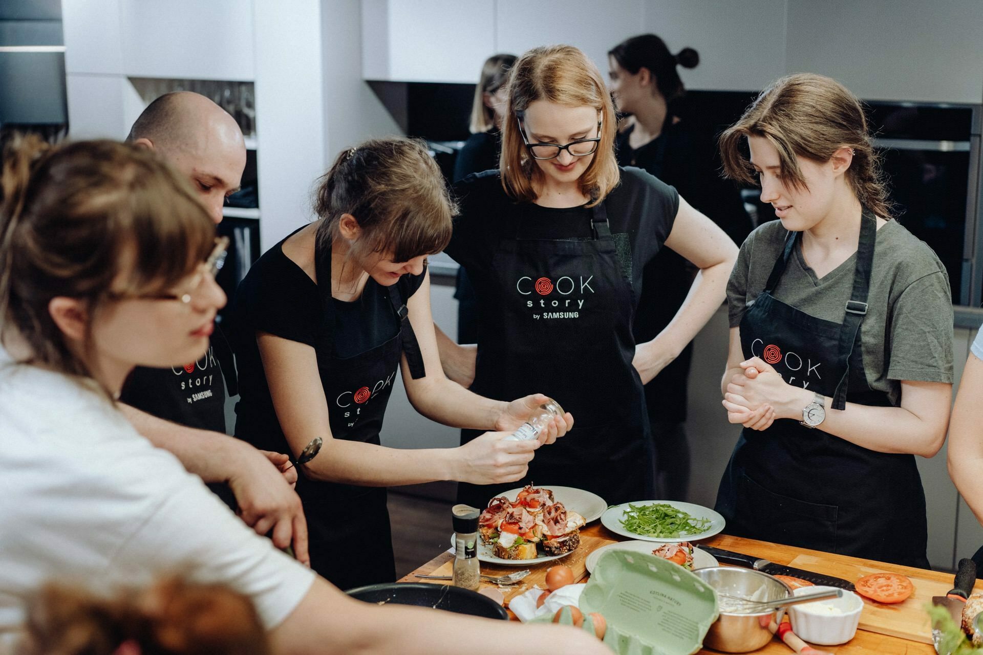 A group of people dressed in black aprons have gathered around the kitchen counter, engaged in cooking. One person pours cream or sauce onto a plate, while others watch and participate in the preparation of various ingredients. The scene captured within the event photography suggests that these are lively cooking classes or workshops.  