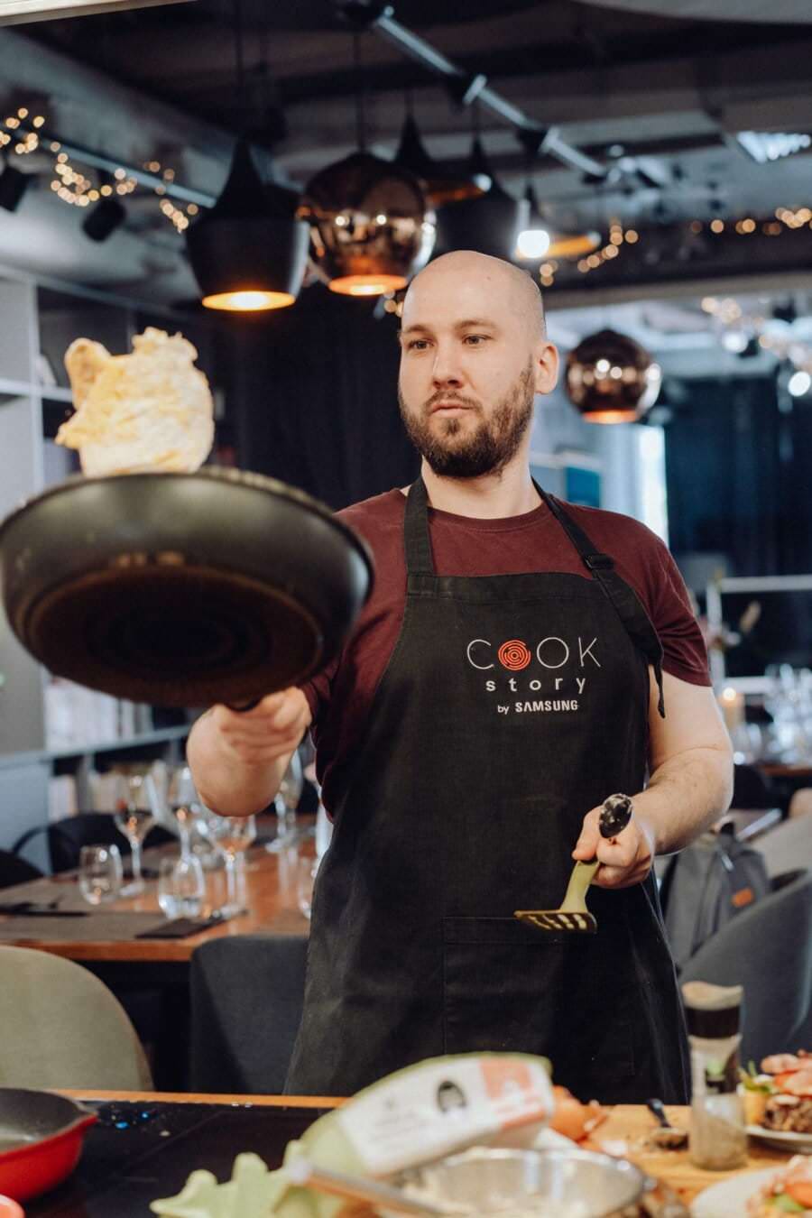 Mężczyzna ubrany w czarny fartuch z napisem „Cook Story by Samsung” przerzuca jedzenie na patelni, trzymając w drugiej ręce szpatułkę. Uchwycony w ramach fotografii eventowej, znajduje się w nowoczesnej kuchni z wiszącymi lampami i stołami widocznymi w tle.