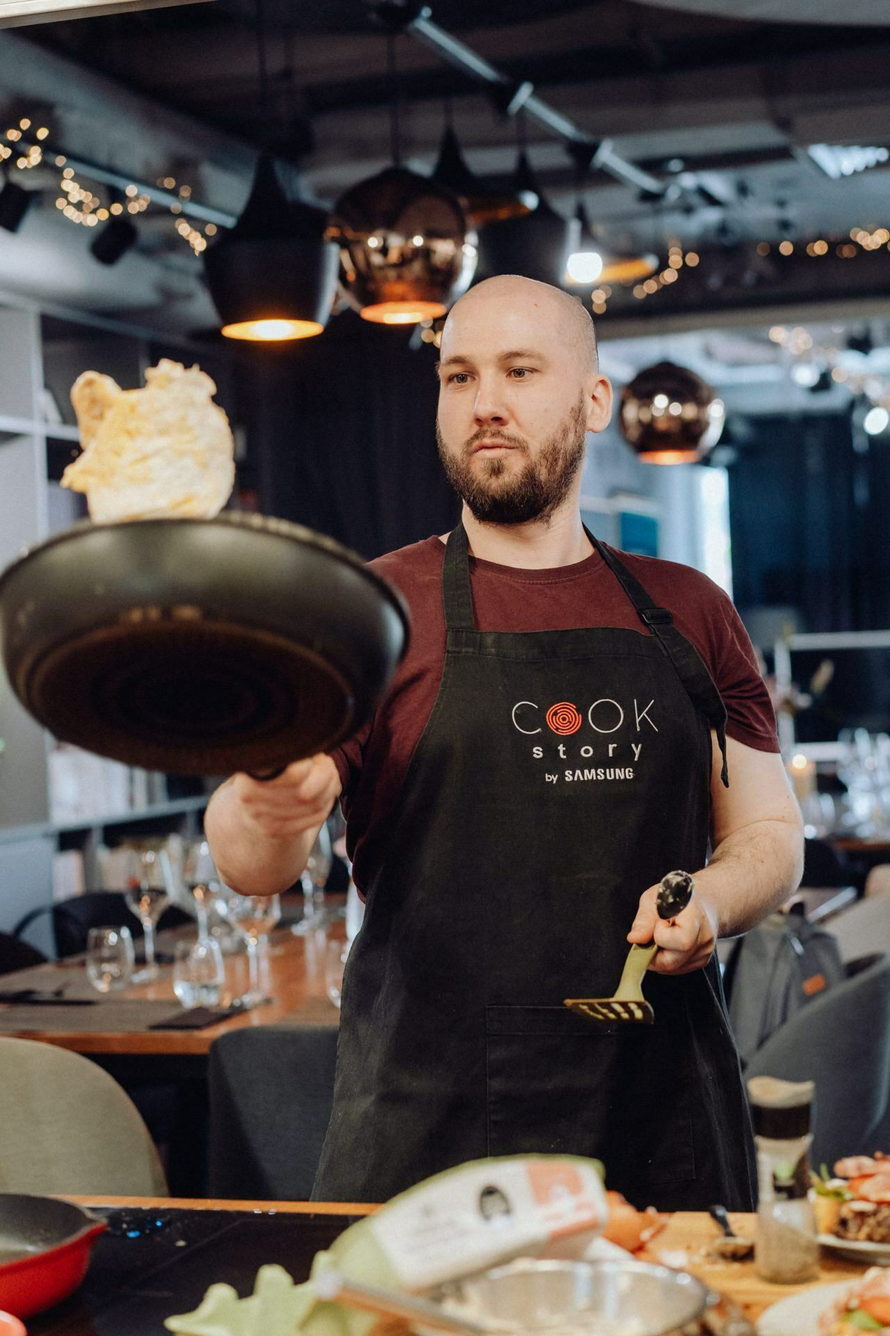 A man dressed in a black apron with the words "Cook Story by Samsung" is flipping food in a pan, holding a spatula in his other hand. Captured as part of an event photograph, he is in a modern kitchen with hanging lamps and tables visible in the background. 