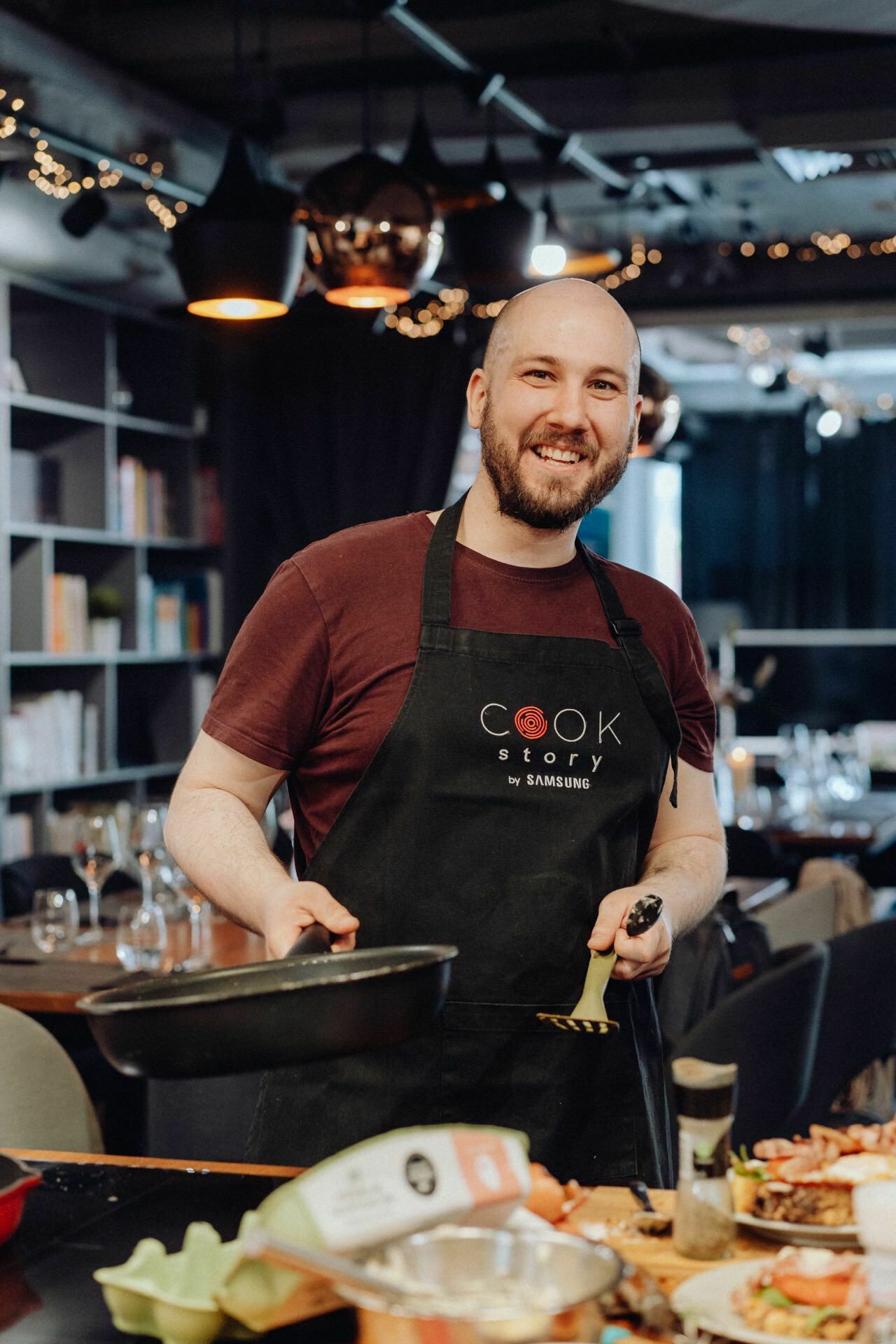 A smiling man with a beard is cooking in the kitchen. He is wearing a black apron with the words "Cook Story by Samsung" and holding a frying pan and spatula. In the foreground, the table is filled with various ingredients and dishes, with bookshelves in the background. It captures the essence of event photography.   