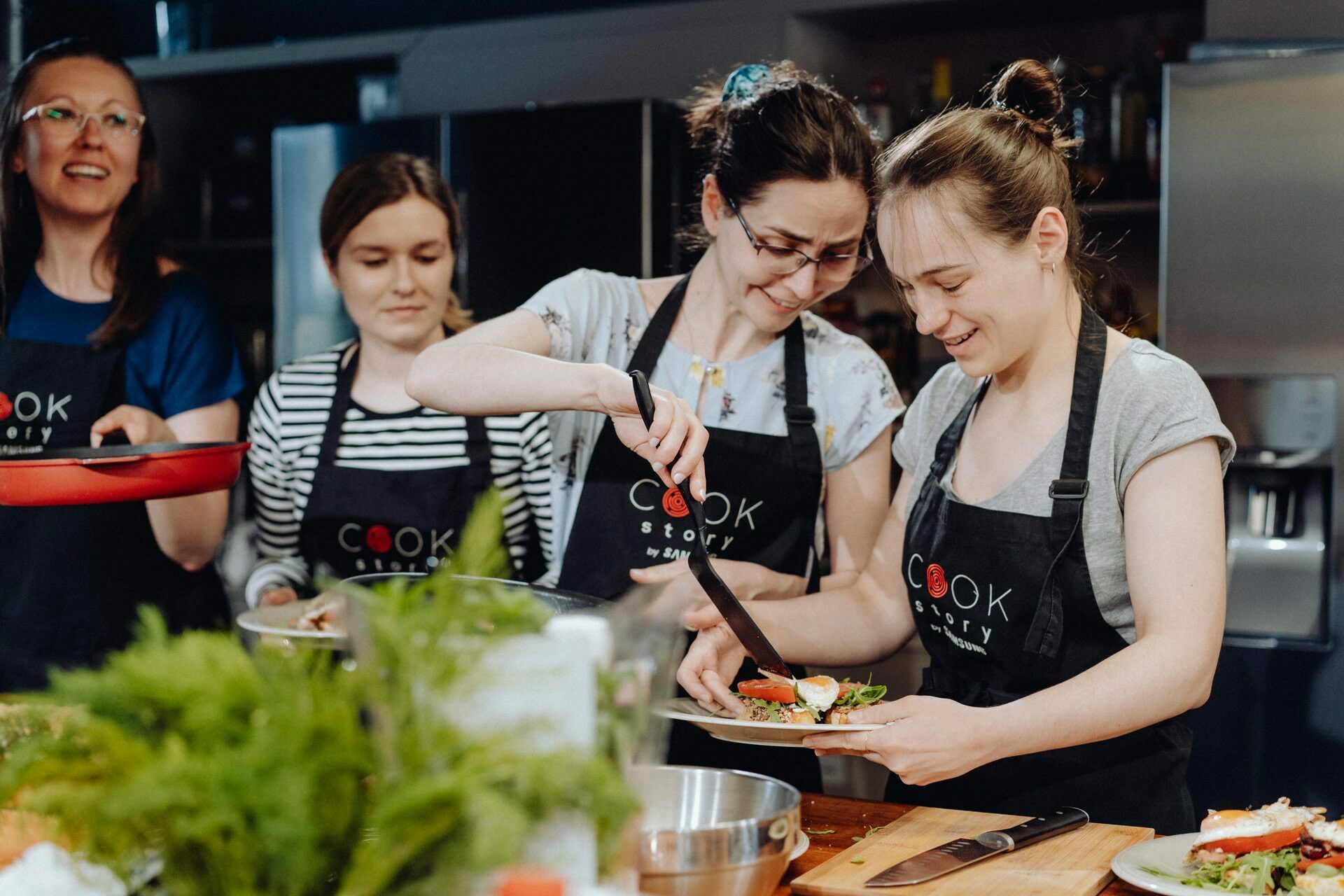 Four women in aprons prepare food in the kitchen. One woman puts decorations on a dish, while another looks on and smiles. The kitchen counter is filled with various ingredients and kitchen utensils. Kitchen appliances and shelves can be seen in the background, capturing the perfect moment for event photography.   