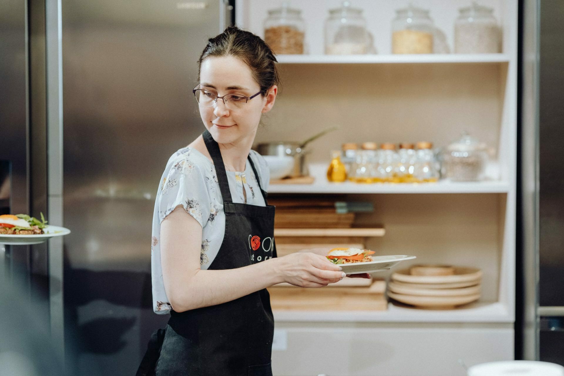 A person wearing glasses and a black apron holds a plate of food in the kitchen. They are standing in front of white shelves filled with jars and dishes, against a backdrop of a countertop and kitchen utensils - a perfect scene for event photography. 