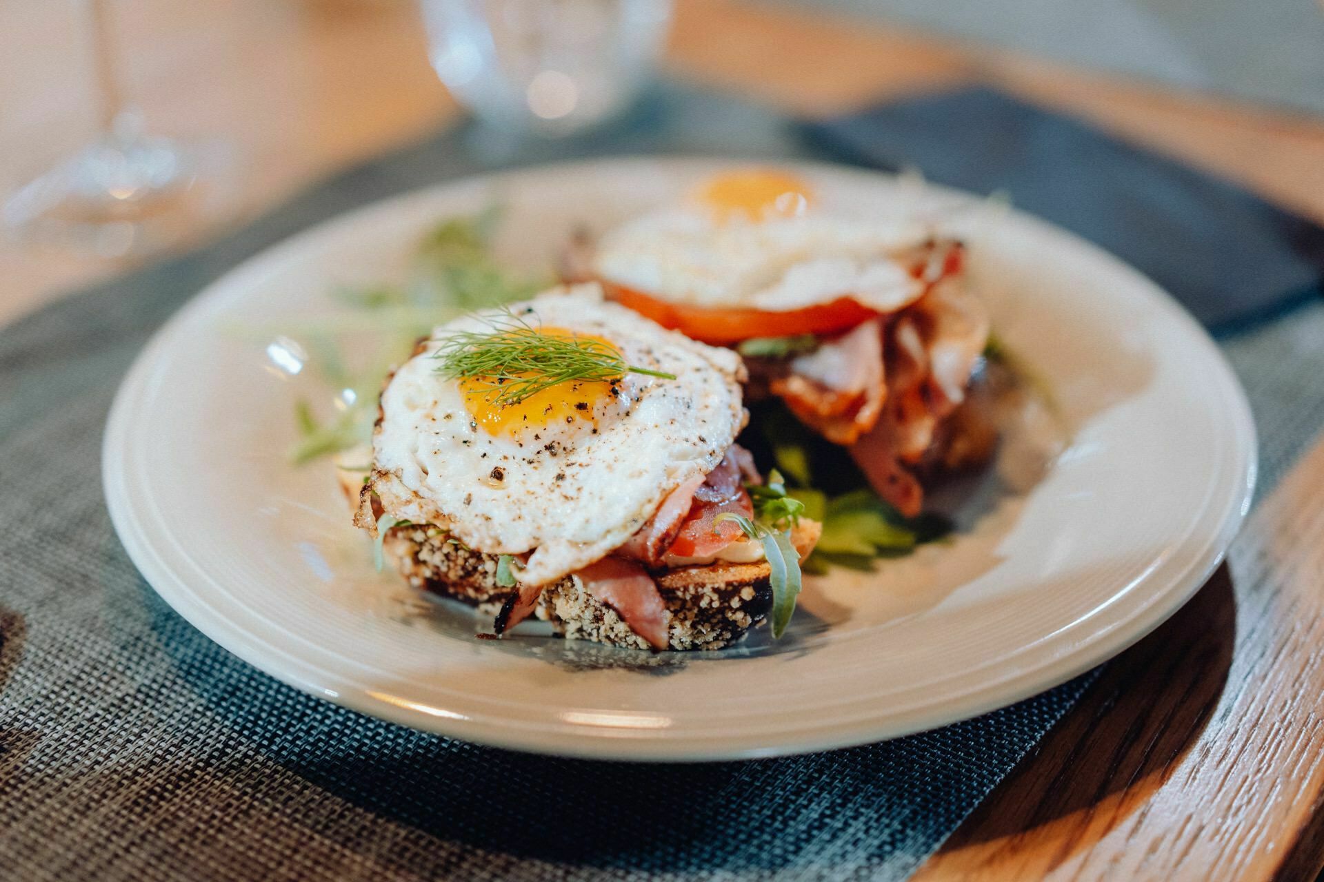 A close-up of an appetizing breakfast dish on a white plate, perfect for an event photography spread. The meal consists of two toasts covered with layers of ham, tomato slices and eggs upside down, decorated with a sprig of fresh dill. The plate is set on a gray placemat on a wooden table.  