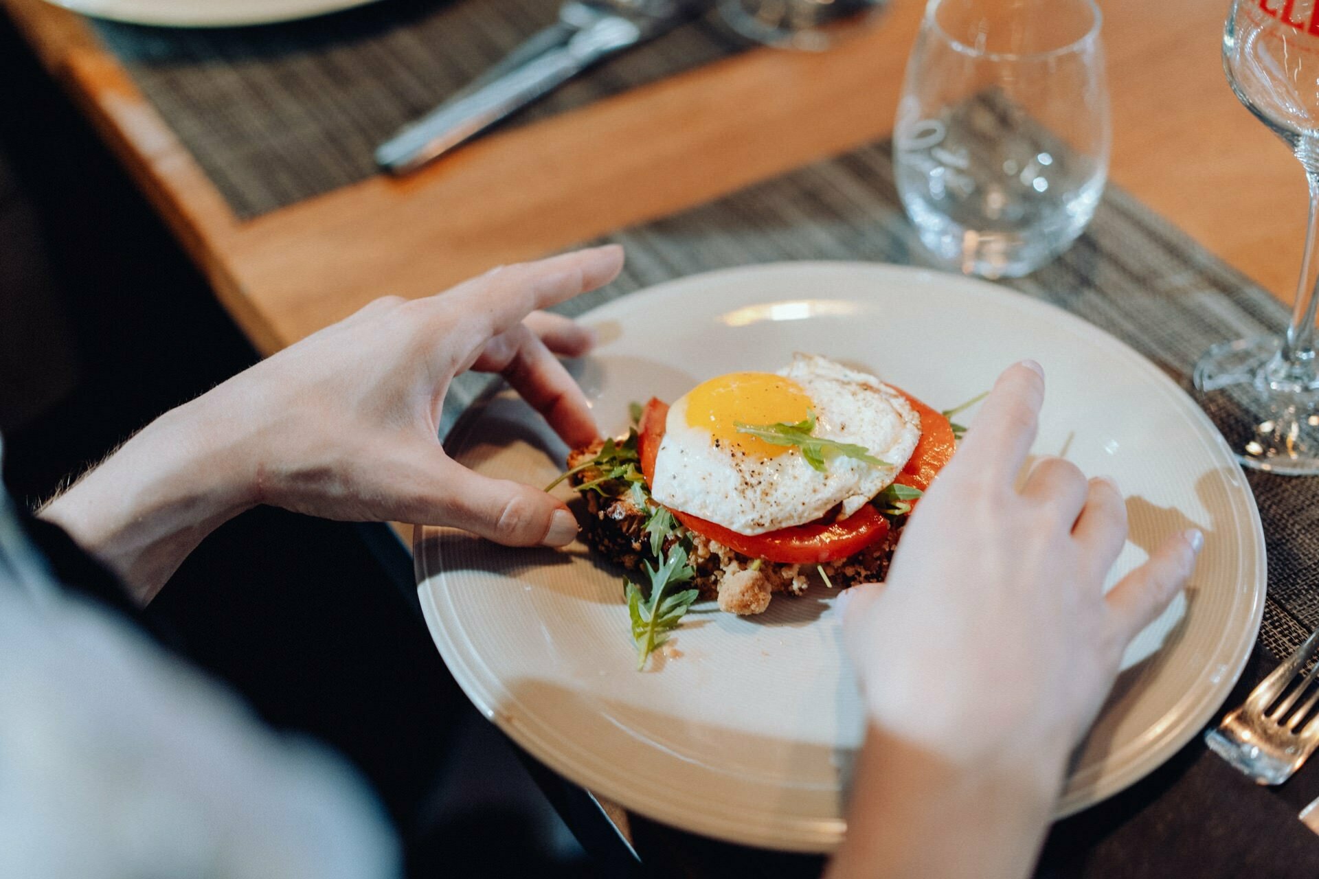 A person places a plate with a dish on a wooden table. The dish consists of an egg sautéed on vegetables, garnished with rosemary. On the table are cutlery, water and wine glasses, which are perfect for capturing in event photography.  