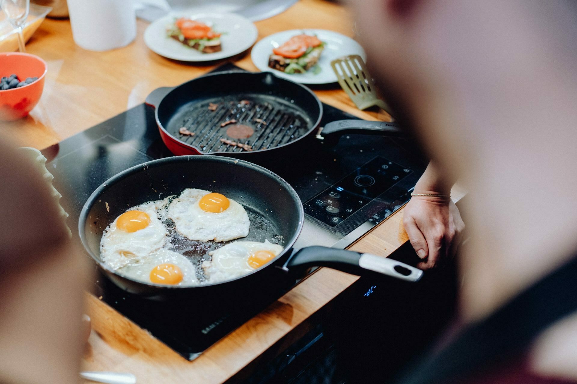 Four eggs are cooked sunny side up in a pan on the electric stove. Next to it is a grill pan with leftover food. Around you can see plates of food, a red bowl and the hands of people preparing food, all set on a wooden countertop - classic scenes captured by event photography in Warsaw.  
