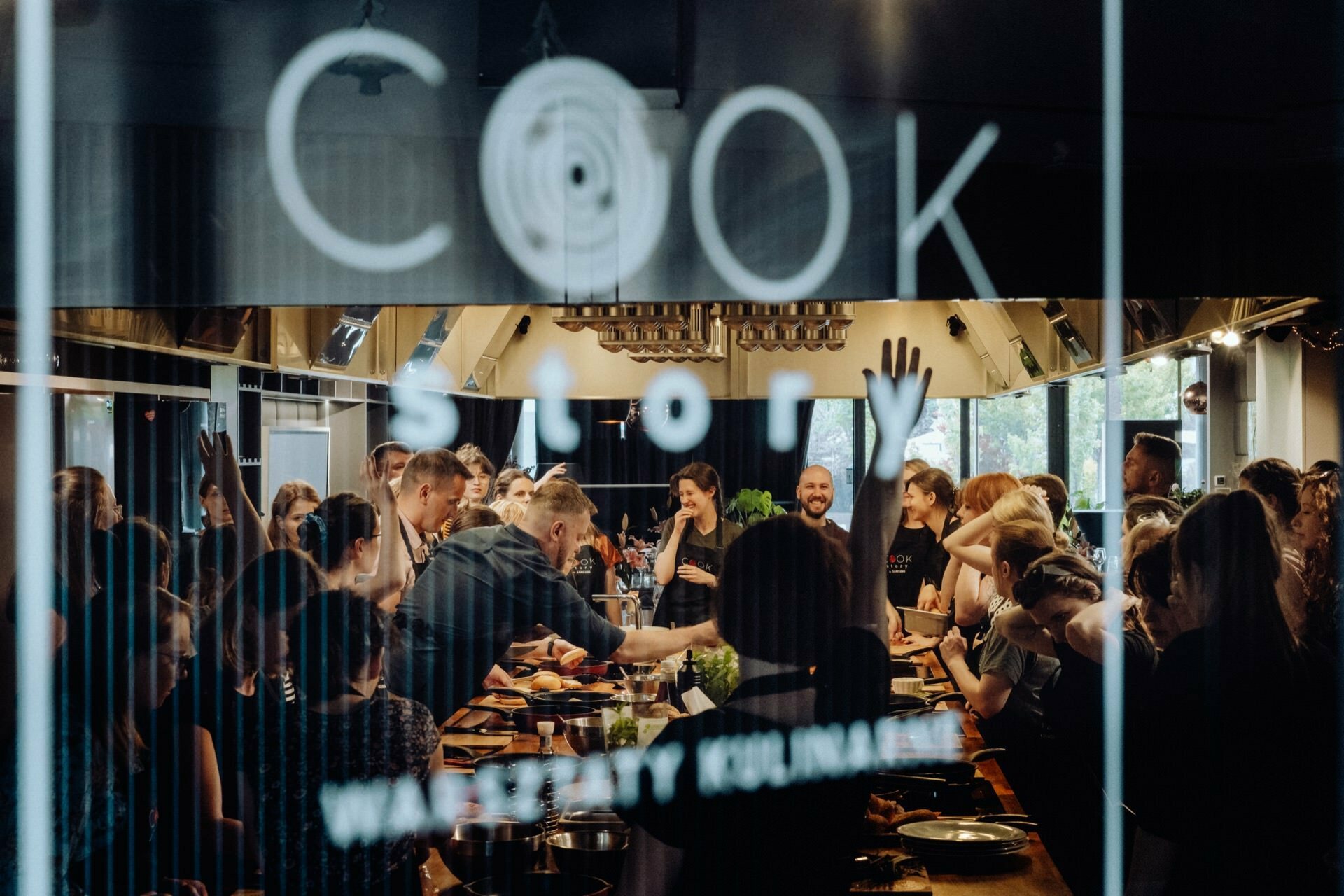 A bustling kitchen filled with people cooking and socializing. The scene is viewed through a glass door that reads "Cook Story," suggesting a cooking workshop or culinary event. The participants seem engaged and enthusiastic - perfect for any event photography that captures vivid moments of culinary joy.  