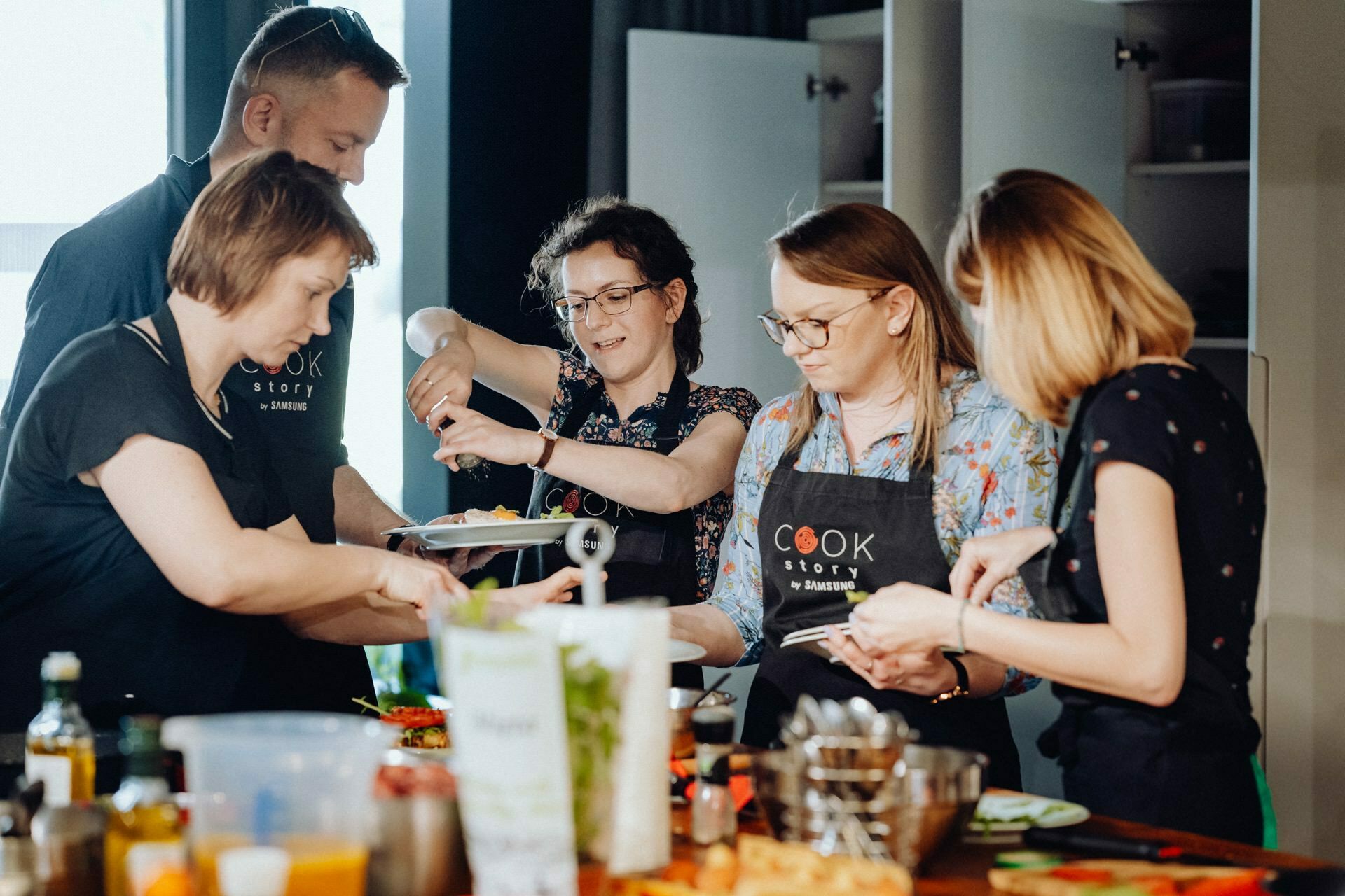 A group of five people dressed in aprons, actively engaged in cooking, gather in the kitchen. They are concentrating on preparing the various ingredients laid out on the table. One person is holding a plate, another is squeezing a spice, and the others are chopping and stirring - a perfect scene for event photography.  