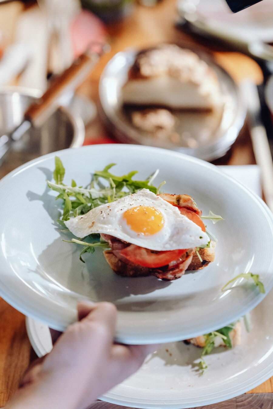 Osoba trzyma biały talerz, na którym leży porcja tostów z jajkiem sadzonym, plasterkami pomidora, awokado i liśćmi rukoli. Na tym zdjęciu eventowym widać kolejny talerz z rozmazanym tłem na drewnianym stole.