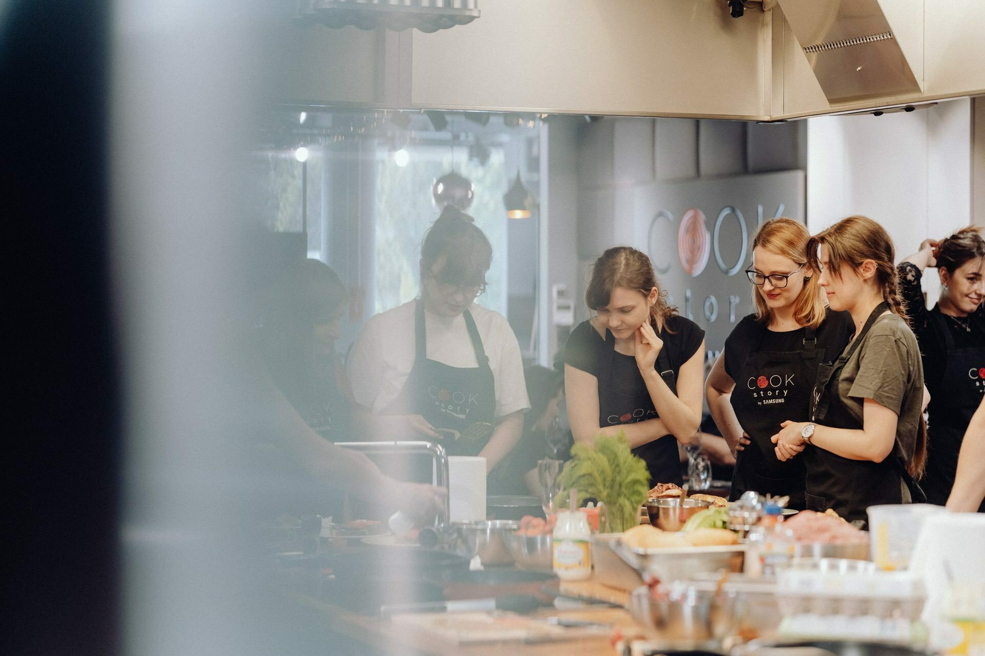 A group of people in aprons stand together in a well-lit kitchen, focused on cooking. In front of them, various ingredients and kitchen tools are spread out on the countertop, creating a friendly atmosphere perfect for event photography. It's like stepping into a vibrant event photo shoot.  