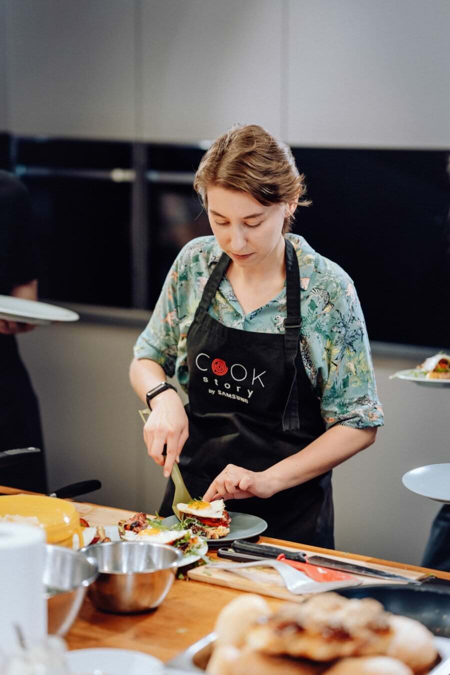 Kobieta ubrana w czarny fartuch z napisem „Cook Story by Samsung” przygotowuje danie w kuchni. Kroi jedzenie na zielonej desce do krojenia ustawionej na drewnianym blacie, otoczona różnymi miskami i składnikami – idealna scena na fotografię eventową.