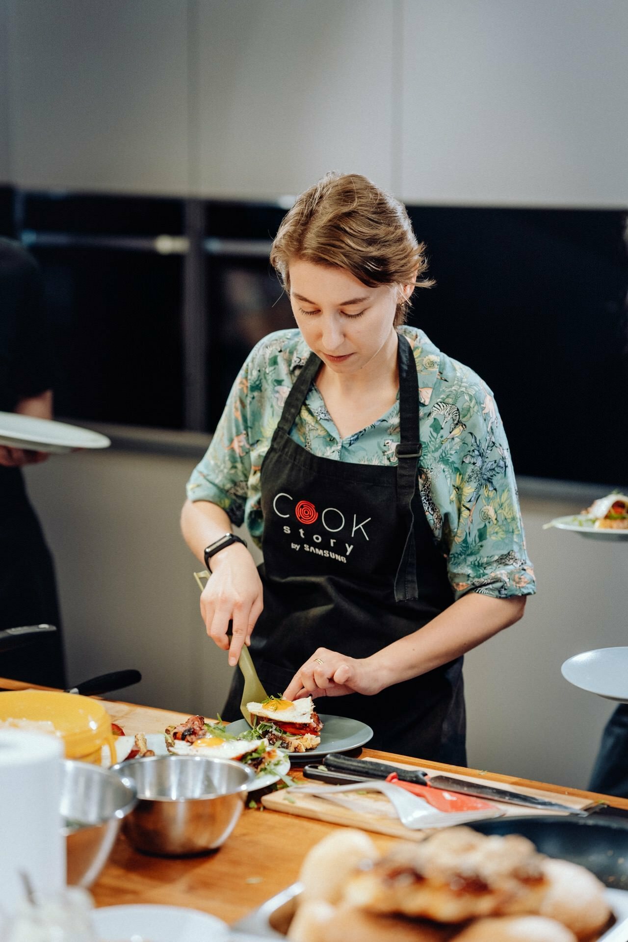 A woman wearing a black apron with the words "Cook Story by Samsung" is preparing a dish in the kitchen. She is cutting food on a green cutting board set on a wooden countertop, surrounded by various bowls and ingredients - a perfect scene for event photography. 