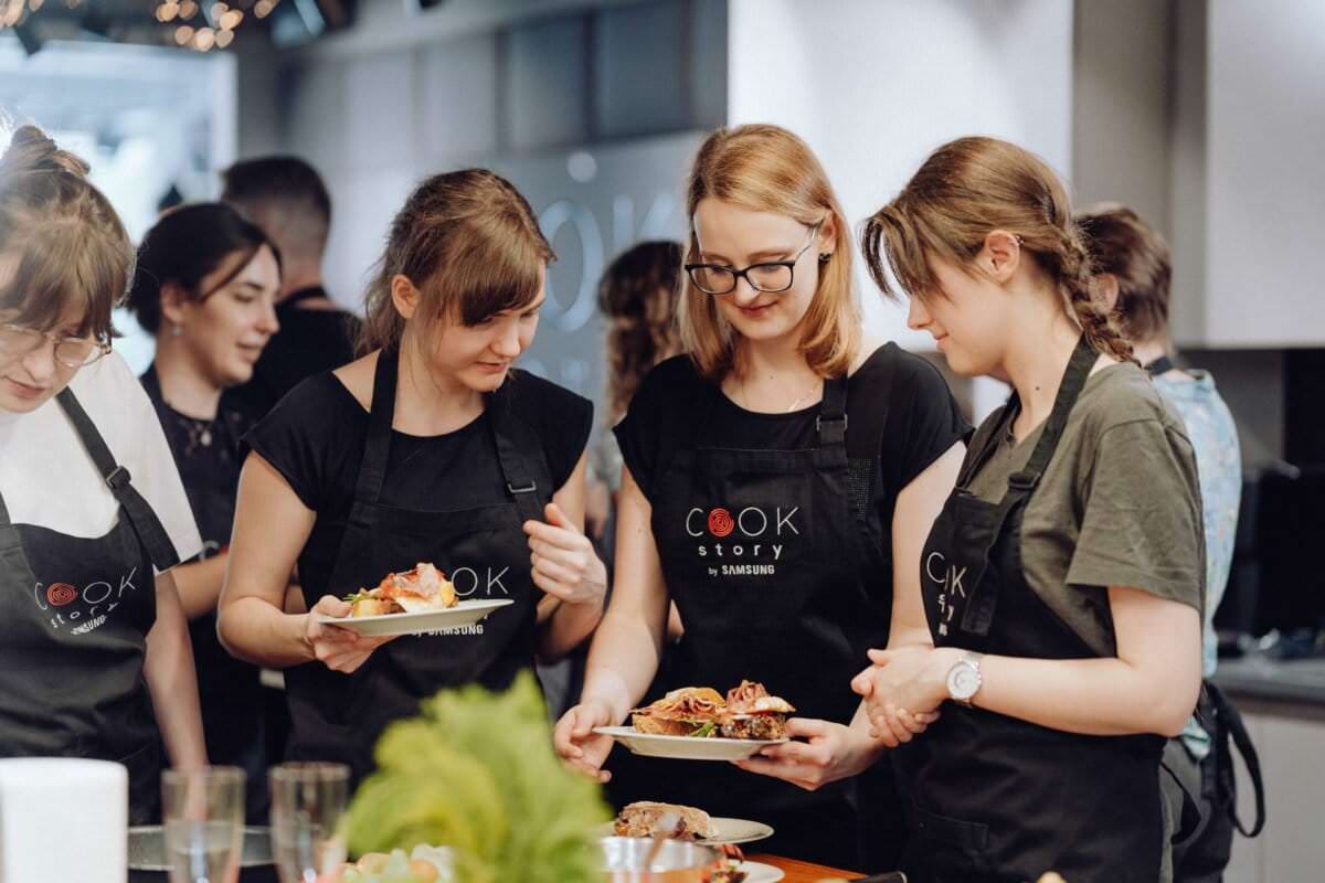 W kuchni gromadzi się grupa osób ubranych w czarne fartuchy z napisem „Cook Story”. Trzy osoby pośrodku trzymają talerze z jedzeniem i prowadzą rozmowę, podczas gdy inni obserwują. W tle sprzęt kuchenny i swobodna atmosfera, przypominająca fotorelację z wydarzeń.