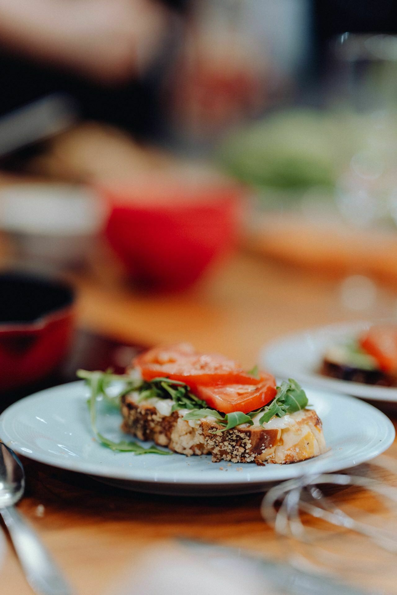 An open-faced sandwich on a white plate sprinkled with arugula, tomato slices and possibly cheese on a slice of seeded bread. In the background you can see a fuzzy table with dishes and red bowls, capturing the moment like an elegant photo-reportage of events. 