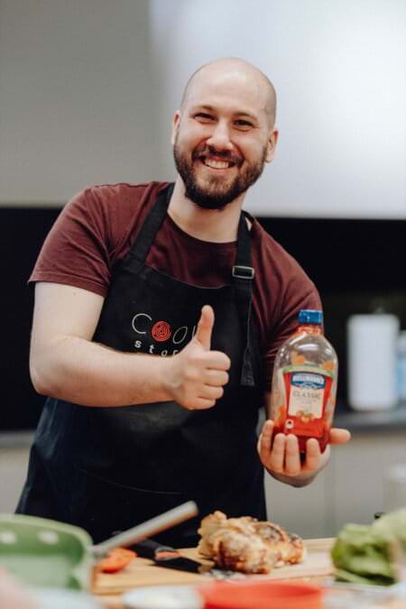 Uśmiechnięty mężczyzna z brodą podnosi kciuki do góry, trzymając butelkę ketchupu Heinz. Ubrany w ciemny fartuch narzucony na bordową koszulkę, stoi w kuchni z różnymi składnikami na blacie przed sobą i ukazuje ten moment w ramach fotografii eventowej.