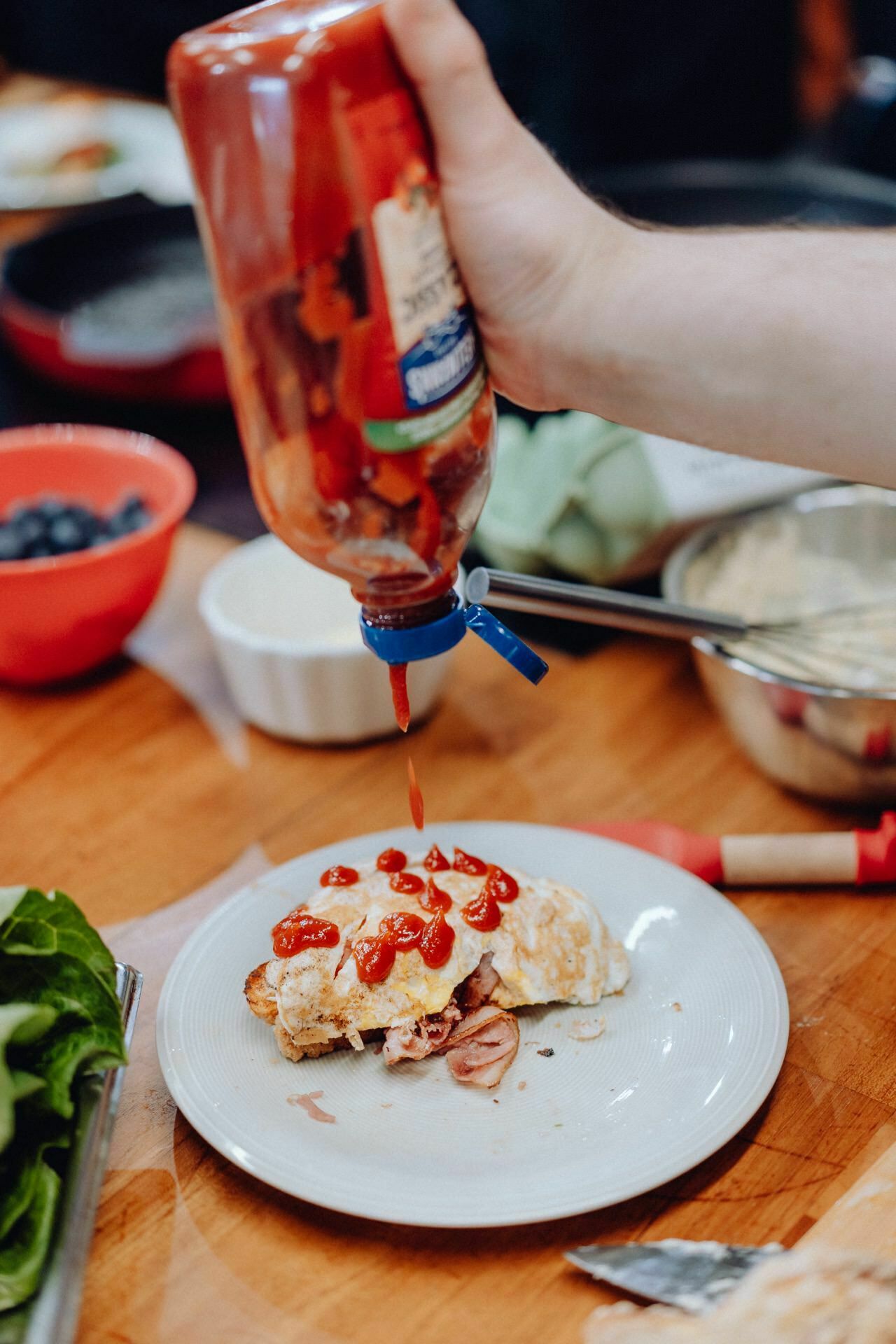 A hand squeezes ketchup from a bottle onto a meal on a white plate. The meal looks like a fried dish with a little sauce. The scene is surrounded by a red bowl of berries, a silver bowl, some lettuce and other kitchen items on a wooden table - perfect for event photography.  