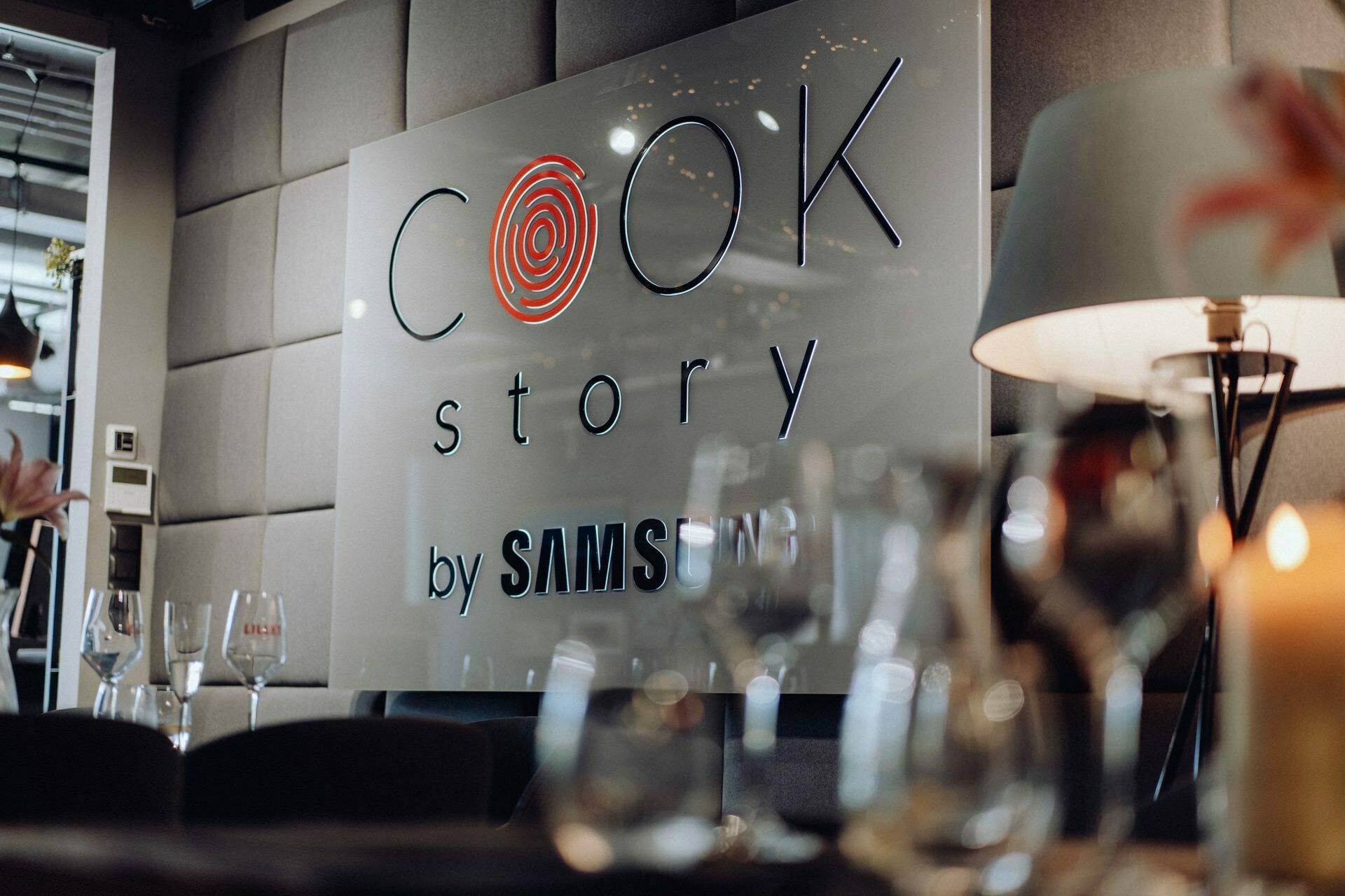 Close-up of a stylish, modern wall sign with the words "COOK Story by SAMSUNG" in a sophisticated font. The background is a padded wall panel in soft shades of gray. In the foreground are fuzzy wine glasses and a lit candle, evoking the exquisite atmosphere of a meal during an elegant event photography.  