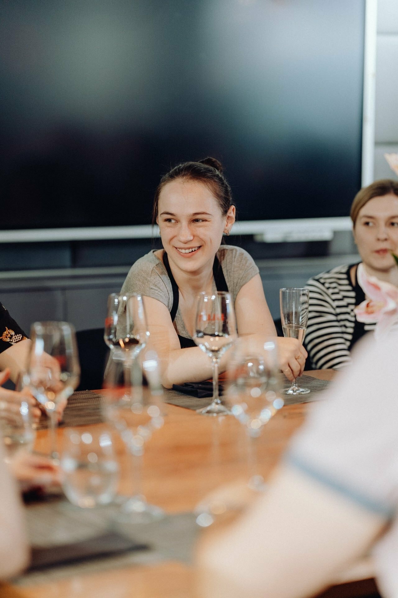 A young woman with her hair tied back sits at a table, smiling and holding a drink in a tall glass. She is surrounded by several empty wine glasses. In the background, other people are sitting at the table, enjoying the casual and social atmosphere - perfectly captured by our event photographer Warsaw.  