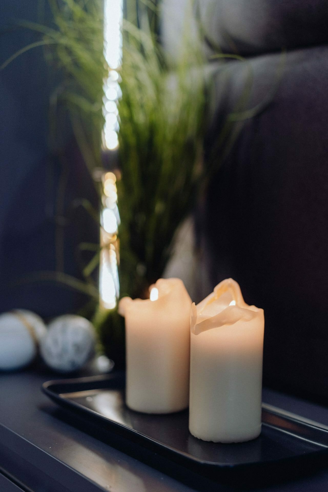 Two partially melted candles stand on a black tray and gently illuminate the surroundings. Behind them, a clump of green grass-like decoration adds a natural element to the dim, cozy surroundings. The background remains out of focus, which enhances the warm glow of the candles, creating a picturesque scene perfect for event photography.  