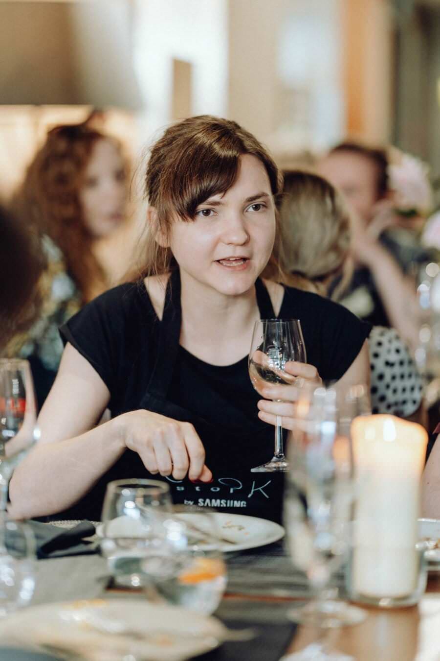 Kobieta o brązowych włosach, ubrana w czarny top i fartuch na szelkach, siedzi przy stole w ruchliwej restauracji, trzymając kieliszek białego wina. Tę scenę można śmiało włączyć do fotorelacji z wydarzeń, która uchwyci ożywioną atmosferę i rozmyte postacie w tle.