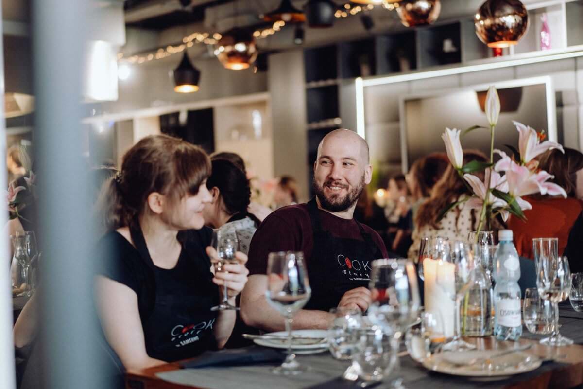 Mężczyzna i kobieta, oboje ubrani w fartuchy, siedzą przy stole i rozmawiają podczas spotkania. Z drinkami w dłoniach, otoczeni są szklankami i kwiatami. Pomieszczenie jest przytulne i dobrze oświetlone, w tle widać inne osoby. Ta urocza scena jest częścią fotorelacji z wydarzenia.