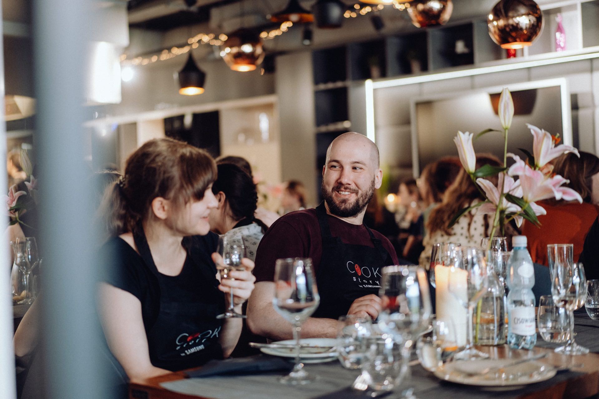 A man and a woman, both dressed in aprons, sit at a table and talk during a meeting. With drinks in their hands, they are surrounded by glasses and flowers. The room is cozy and well-lit, with other people visible in the background. This charming scene is part of a photo report of the event.   