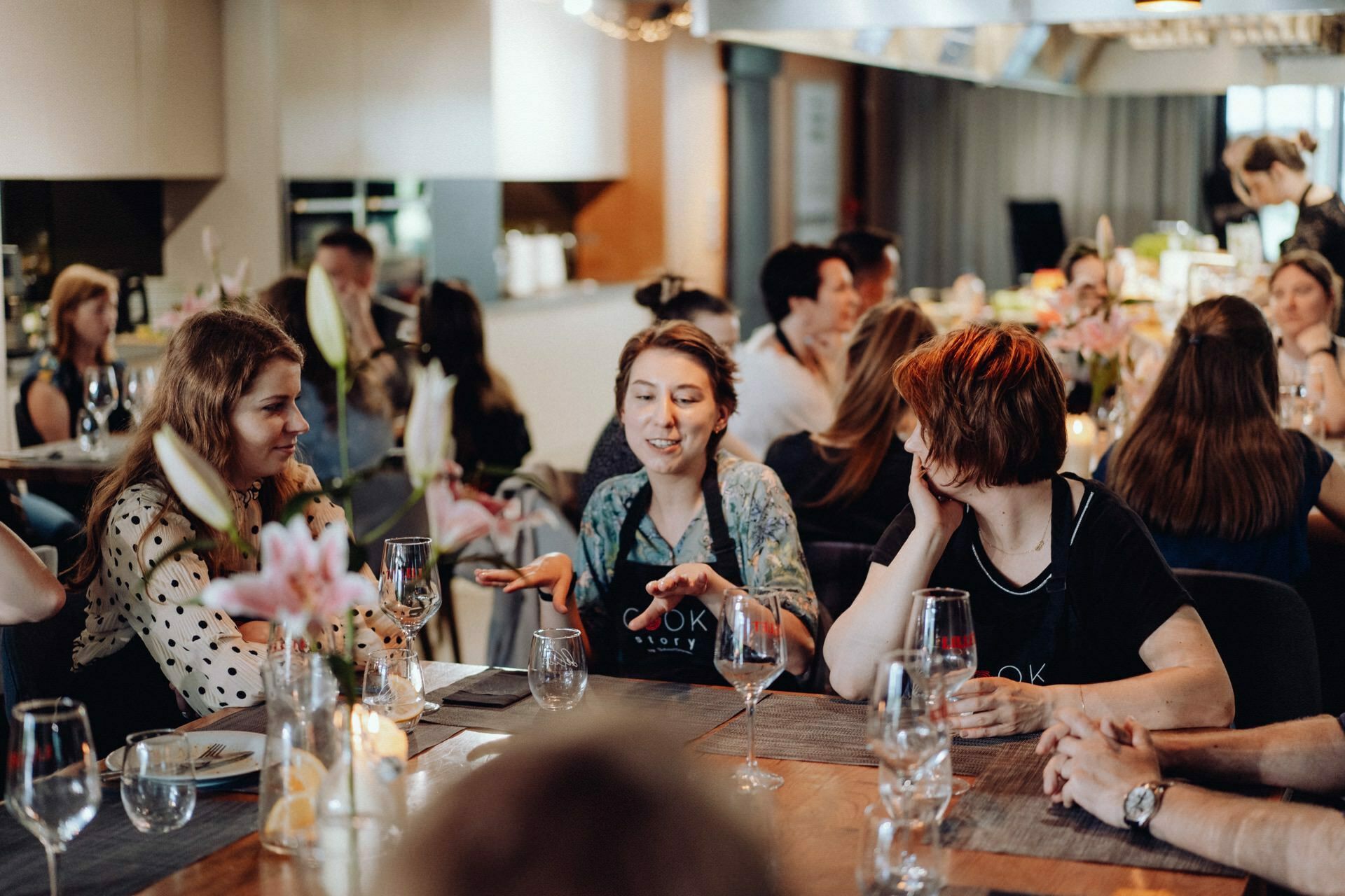 A group of people sit around a table during a lively social gathering. In the foreground, three women are talking and one of them is gesticulating. The table is set with wine glasses, flowers and cutlery. A perfect scene for event photography captured by event photographer Warsaw.   