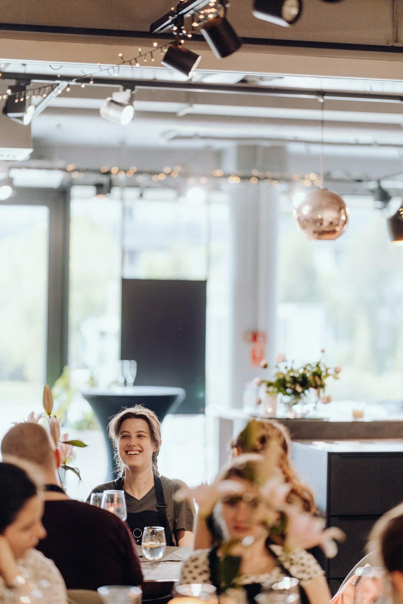 A young woman sits at a table with a broad smile, holding a glass. She is in a modern, well-lit room, decorated with fairy lights and reflective decorations in the background. Around her sit several other people engaged in conversation - capturing the essence of event photography.  