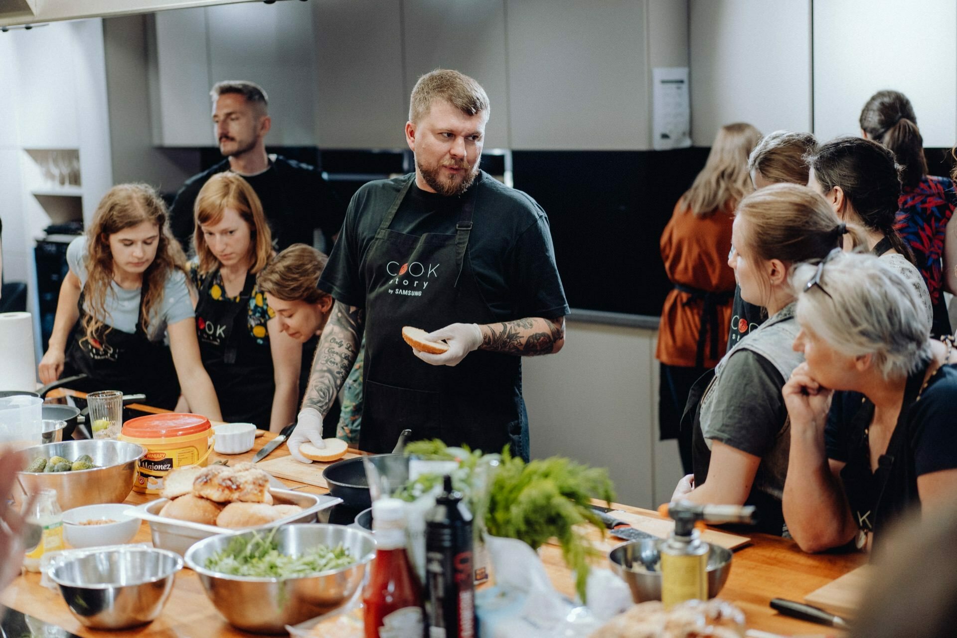 A group of people carefully follow a bearded man with tattoos as he demonstrates cooking techniques at a large wooden table. While participants watch and take notes, various ingredients and cooking utensils are laid out, creating an immersive photo essay of the events. 