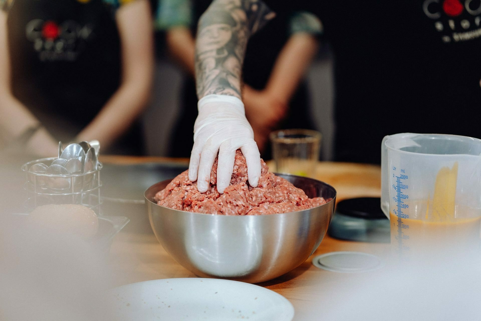 A person wearing a white glove is squeezing minced meat in a metal bowl on a wooden table, as part of what could be an interesting photo-report of events. Other cooking utensils and ingredients are partially visible around the table, with another person standing in the background. 