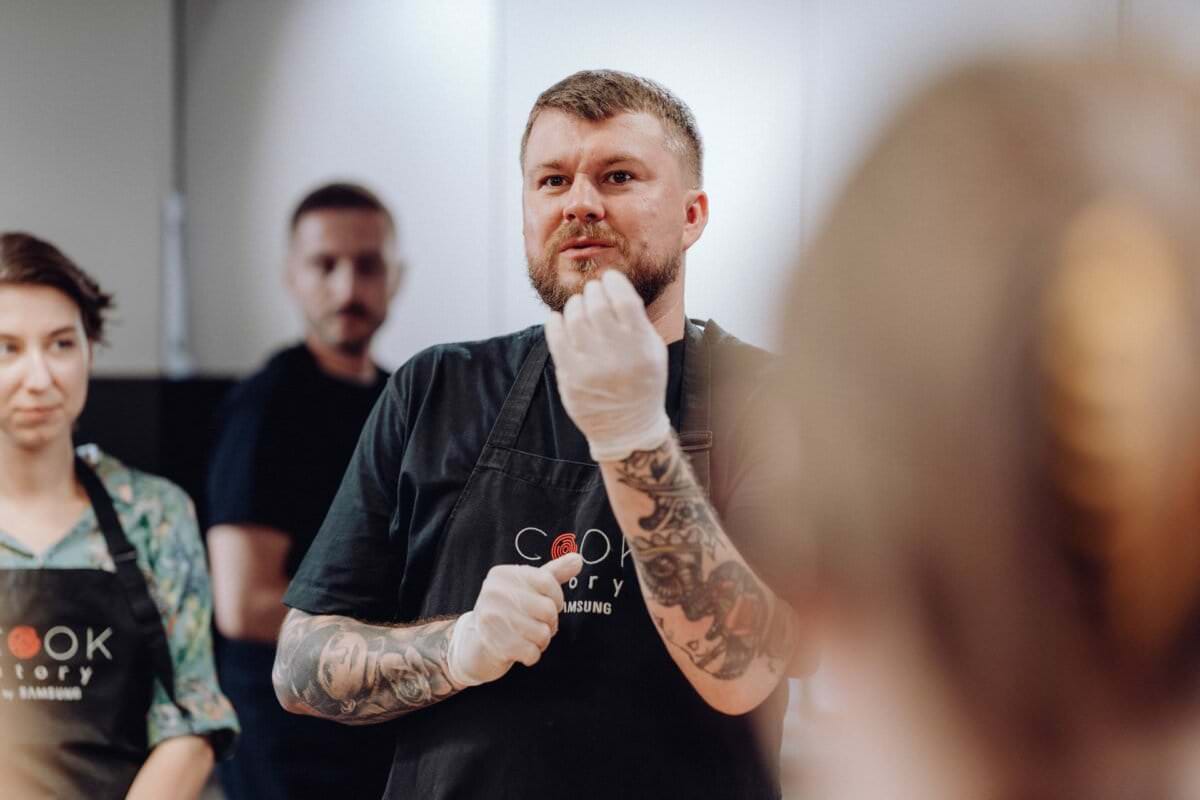 Z pasją mówi mężczyzna z tatuażami, ubrany w czarny fartuch i białe rękawiczki, gestykulując rękami. W tle częściowo widoczne są dwie inne osoby w fartuchach, które uważnie go przysłuchują w pomieszczeniu idealnym do robienia zdjęć eventowych.