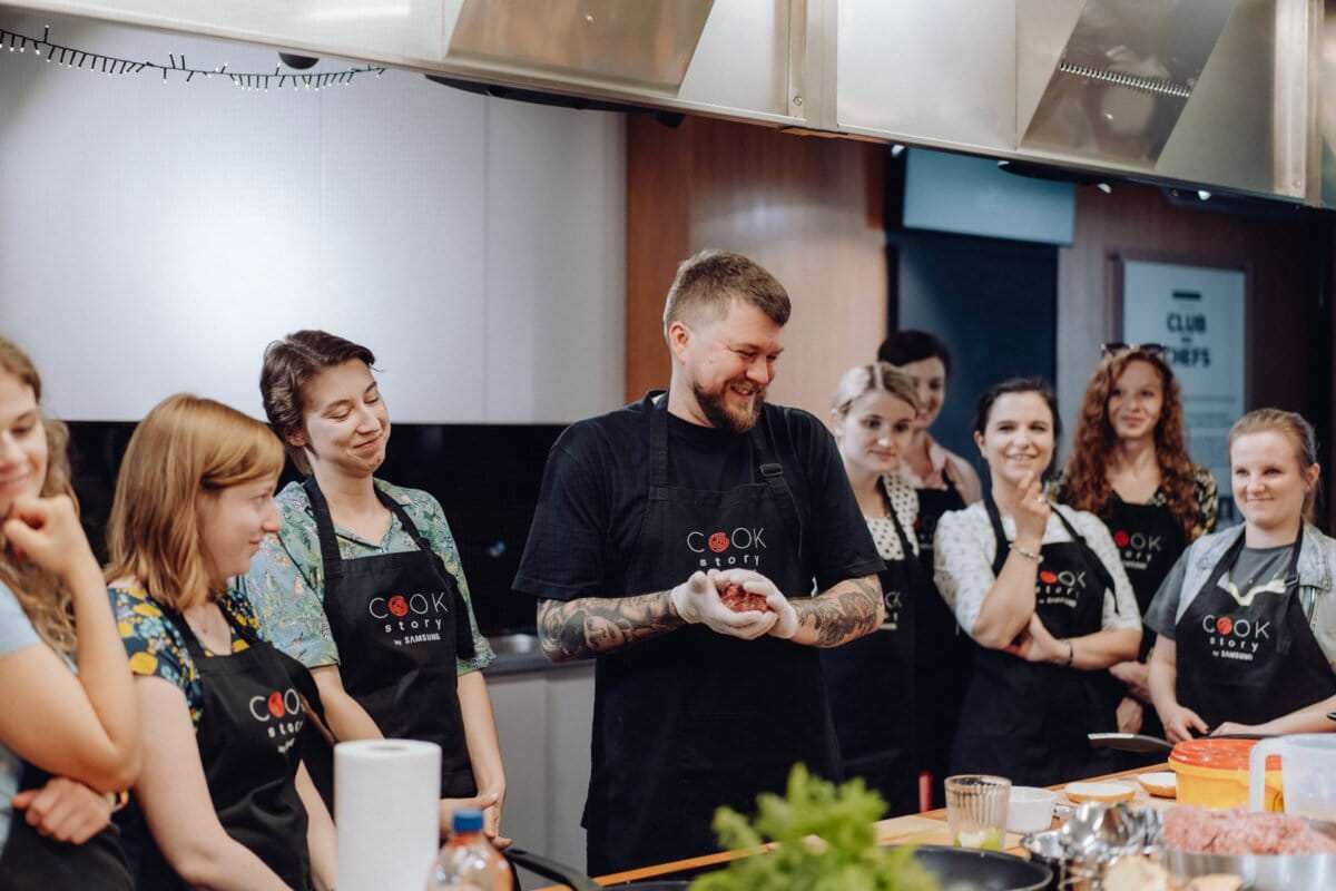 Grupa osób w fartuchach stoi wokół blatu kuchennego podczas lekcji gotowania. Instruktor, uśmiechnięty i rozmawiający, stoi pośrodku, a uczestnicy uważnie się przyglądają. Na blacie rozłożone są różne składniki i przybory kuchenne, tworząc idealne zdjęcie eventowe, które uchwyci tętniącą życiem atmosferę.