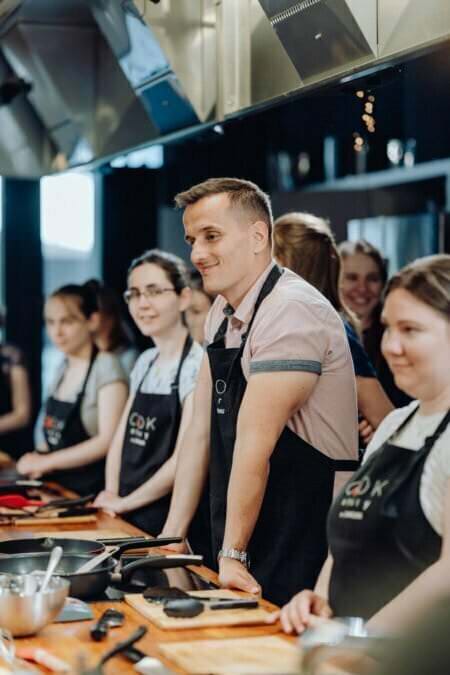 Grupa osób ubranych w czarne fartuchy stoi wokół blatu kuchennego z przyborami kuchennymi. Jeden mężczyzna w jasnoróżowej koszuli stoi w widocznym miejscu pośrodku i uśmiecha się. Wygląda na to, że biorą udział w lekcjach gotowania, pięknie uchwyconych na potrzeby sesji fotograficznej eventowej.