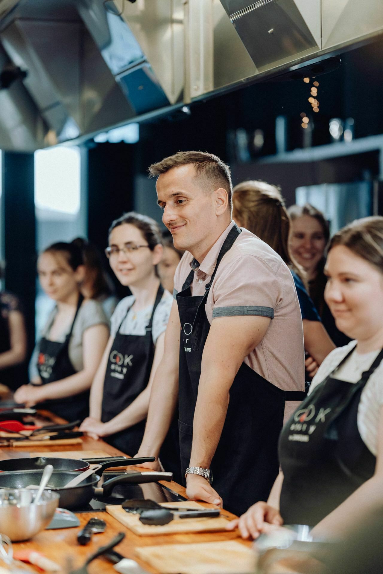 A group of people dressed in black aprons stand around a kitchen counter with kitchen utensils. One man in a bright pink shirt stands prominently in the middle and smiles. They appear to be taking part in a cooking class, beautifully captured for the event photo shoot.  