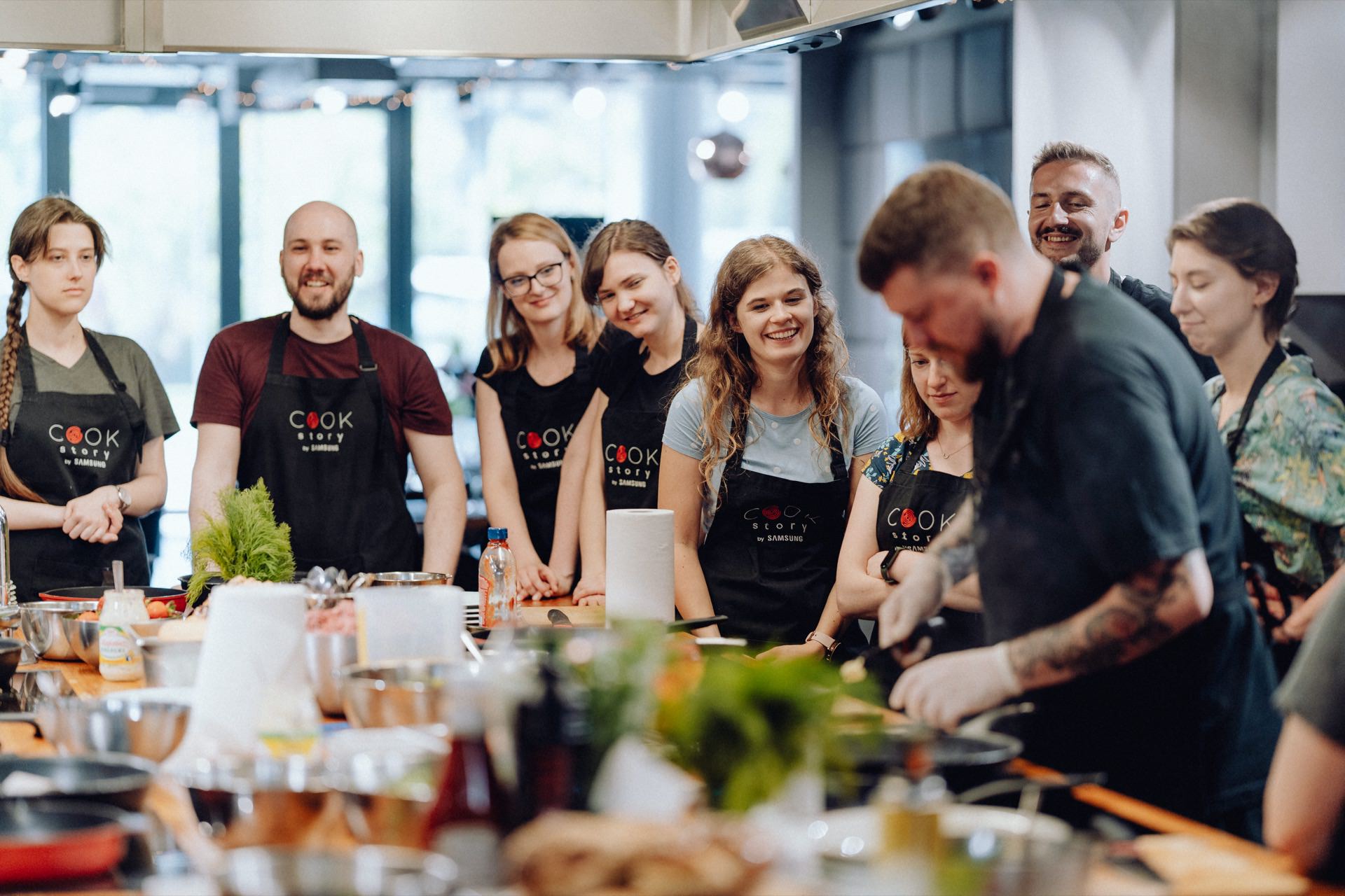 A group of people dressed in aprons gather around a kitchen station, watching a man as he cooks. The station is filled with various ingredients and utensils. They look engaged and happy, suggesting that this could be a cooking lesson or demonstration in a well-lit kitchen - an ideal scene for event photography.  