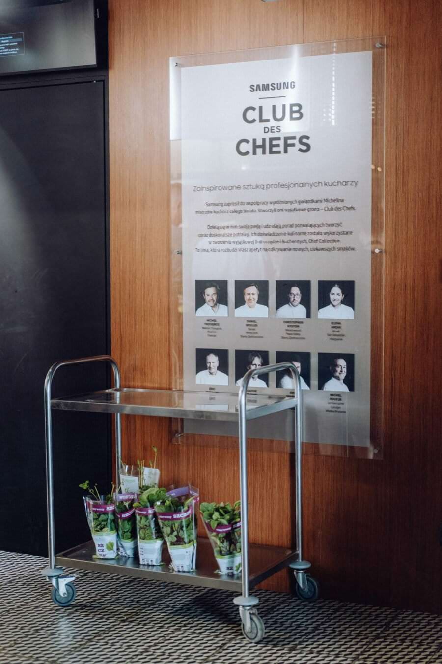 Metalowy wózek z kilkoma doniczkowymi roślinami ziołowymi stoi obok drewnianej ściany ozdobionej plakatem przedstawiającym „Samsung Club des Chefs” ze zdjęciami i nazwiskami szefów kuchni. Podłoga ma wzór w kratkę. Tekst na plakacie jest w języku polskim, co czyni go idealnym miejscem na fotografię eventową.