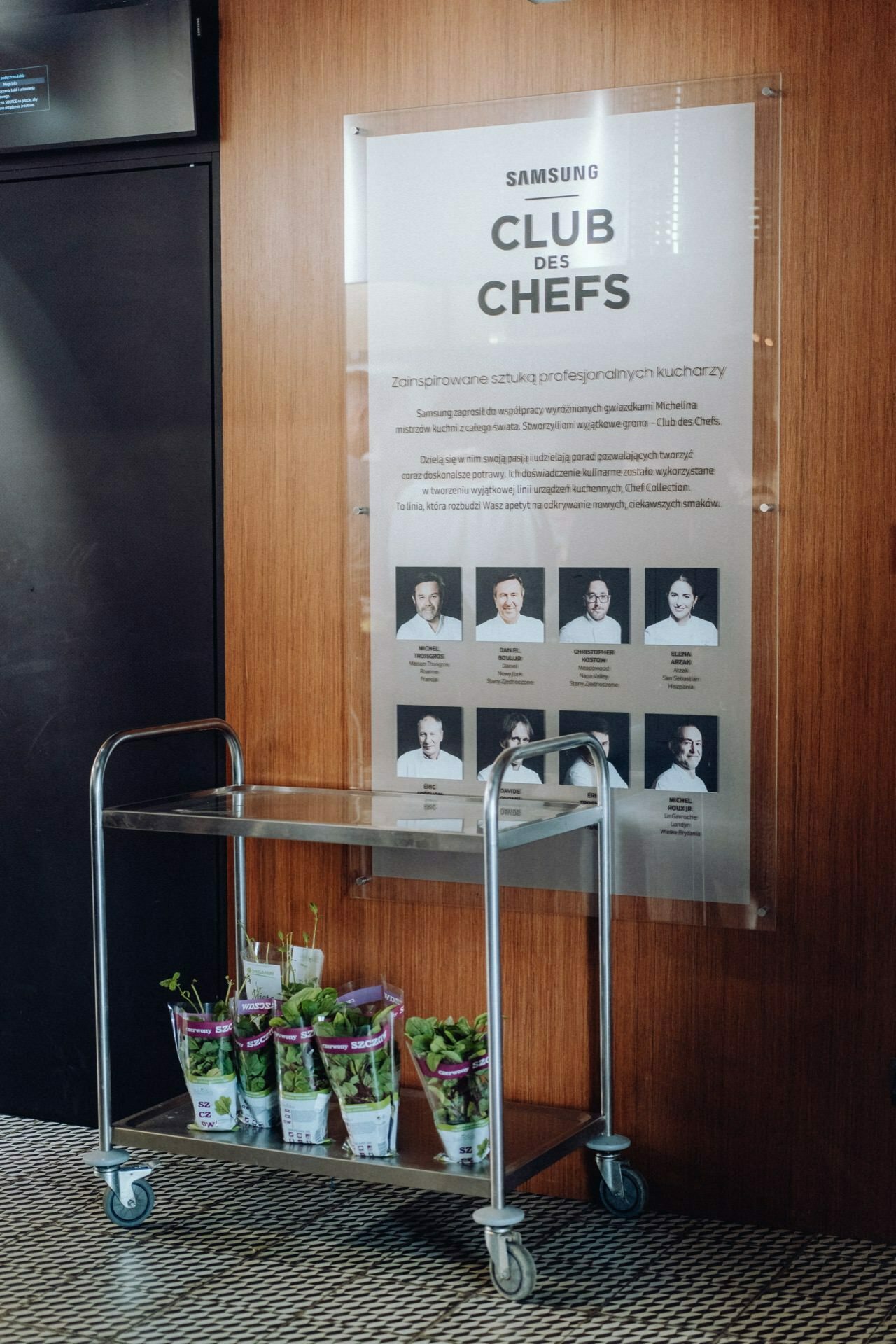 A metal cart with several potted herb plants stands next to a wooden wall decorated with a poster depicting the "Samsung Club des Chefs" with photos and names of chefs. The floor has a checkered pattern. The text on the poster is in Polish, making it ideal for event photography.  