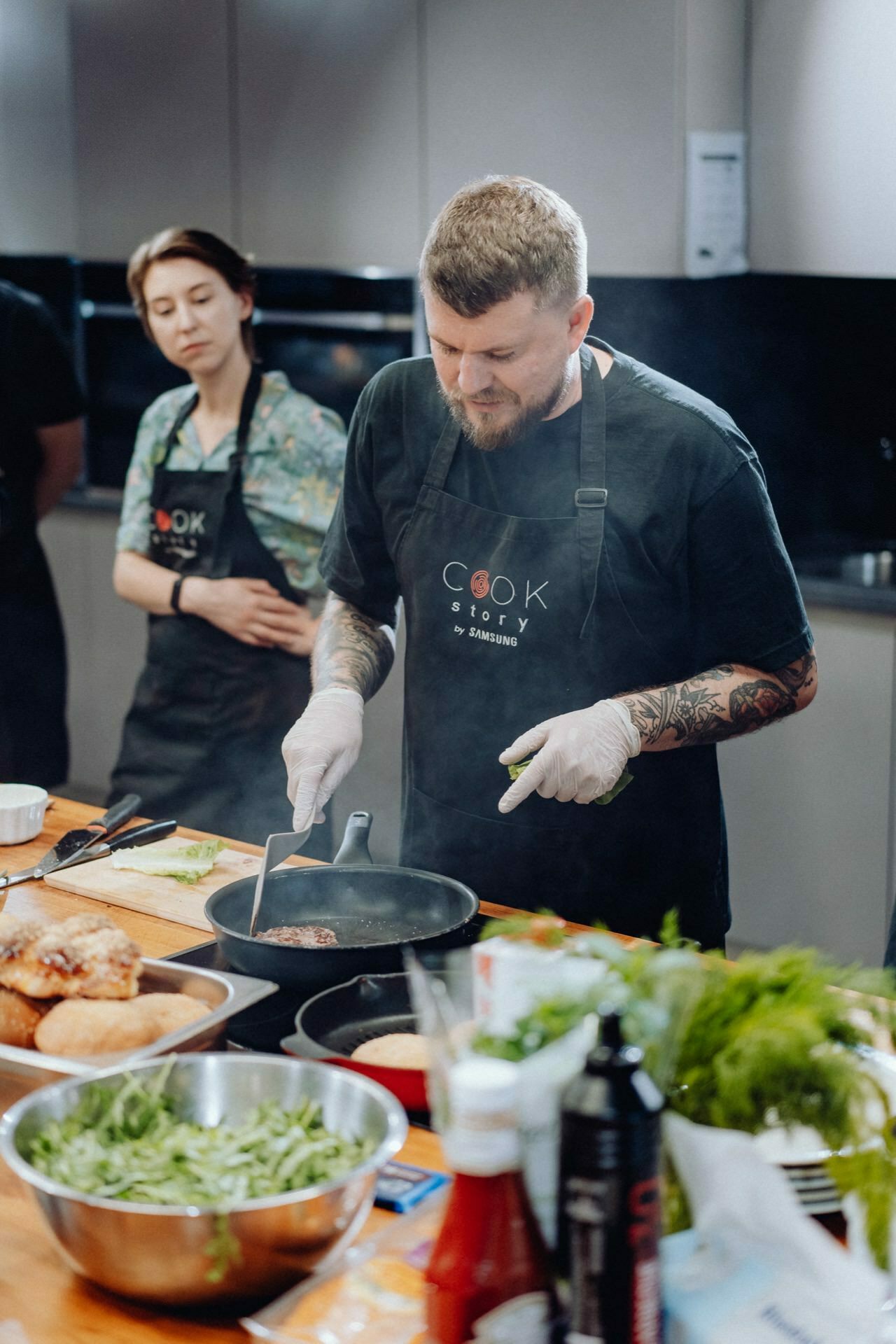 A bearded man dressed in a black apron and gloves is cooking in the kitchen while a woman, also wearing an apron, watches. The man is focused on the sizzling pan in front of him. On the countertop are various ingredients, including vegetables and sandwiches - perfect moments for event photography.  