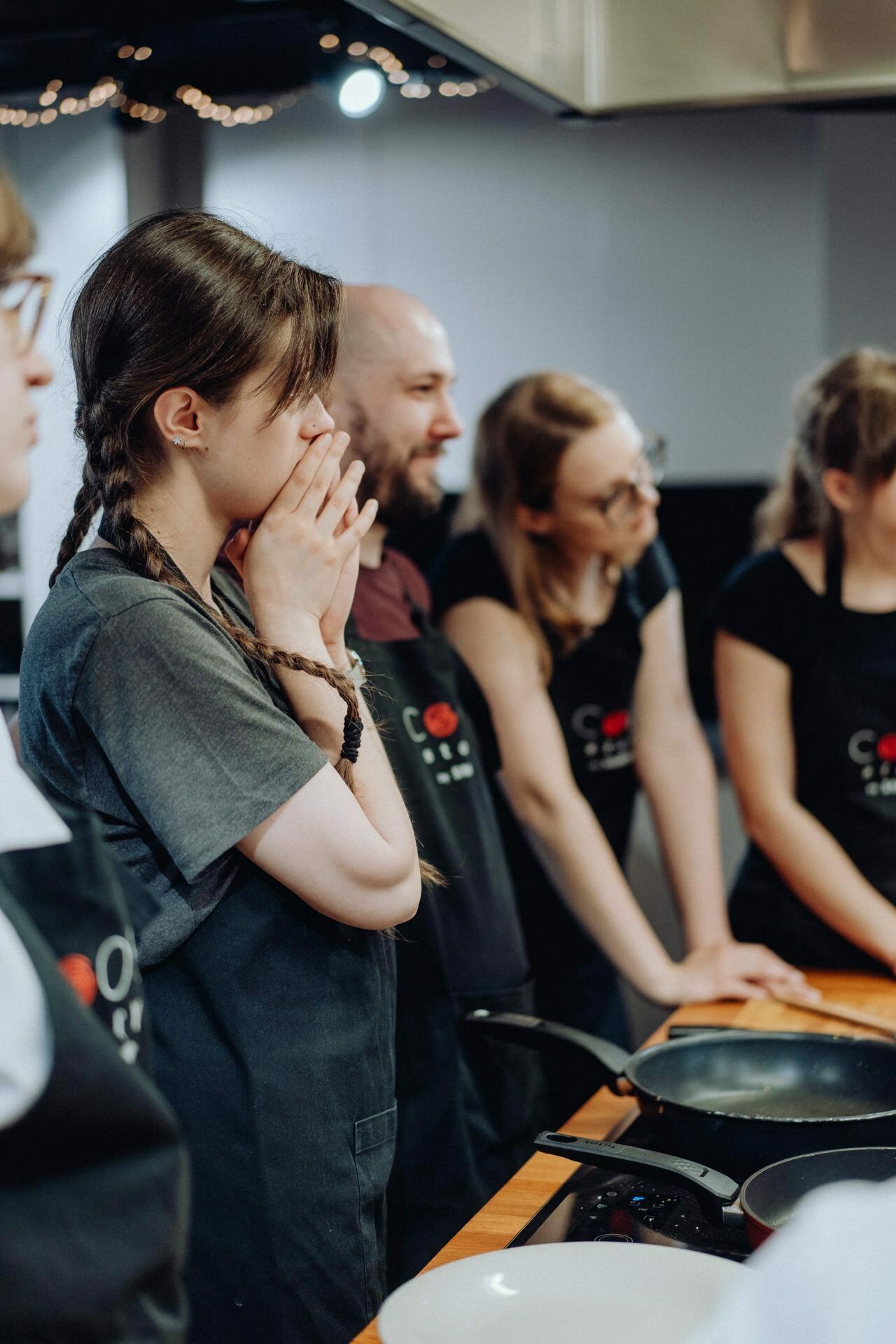 A group of people dressed in black aprons stand around the cooking station. In the foreground, a woman with braids covers her mouth with her hands and looks concerned. Others are focused on the cooking process, with pans and ingredients on the countertop in front of them, as captured perfectly by the photo report of the events.  