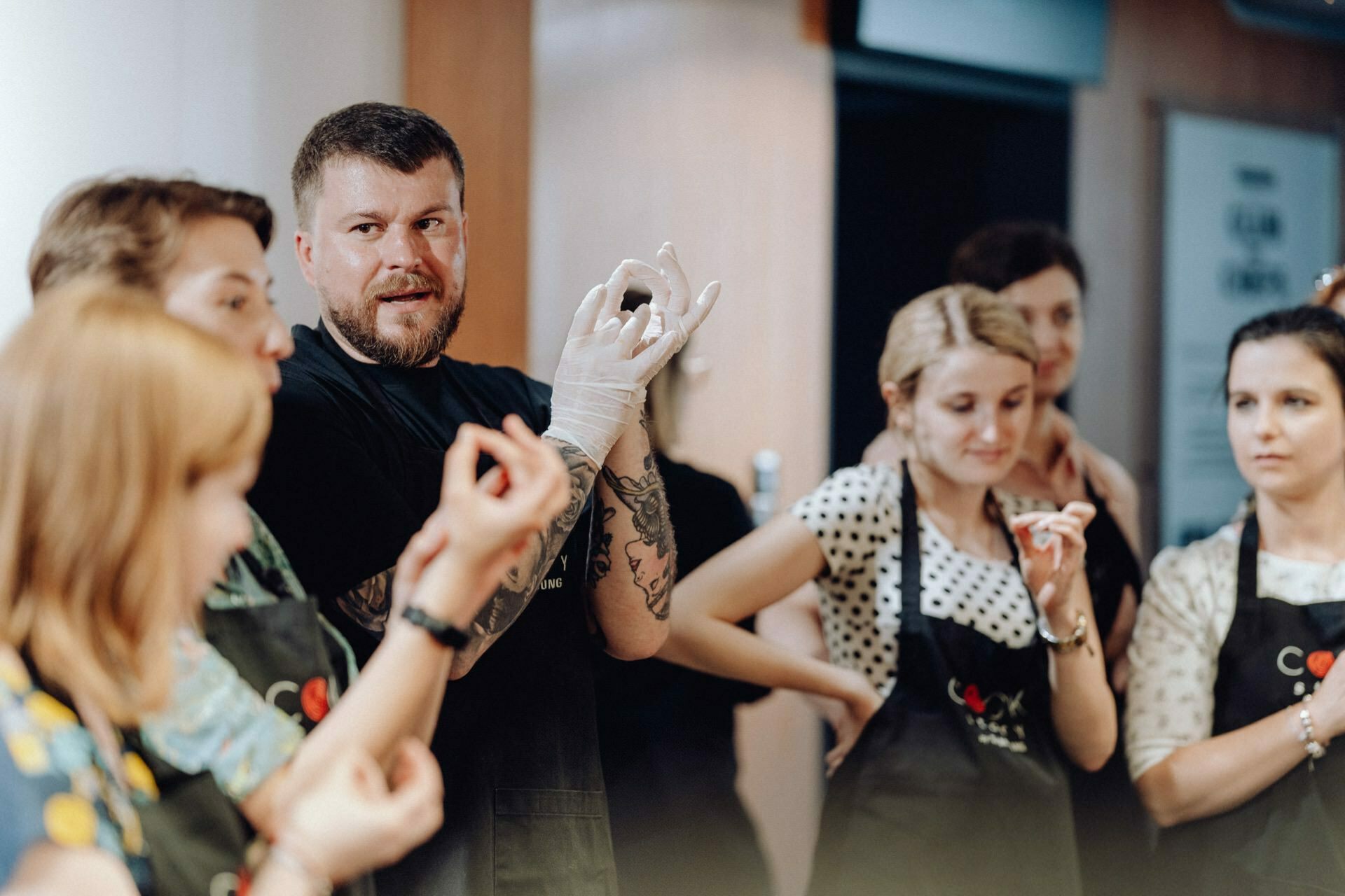 A man wearing gloves and gesturing with his hands leads a cooking class, explaining the technique to a group of attentive participants. The group, consisting mainly of women in aprons, stands together in the kitchen, perfect for a photo shoot that captures the essence of the workshop. 