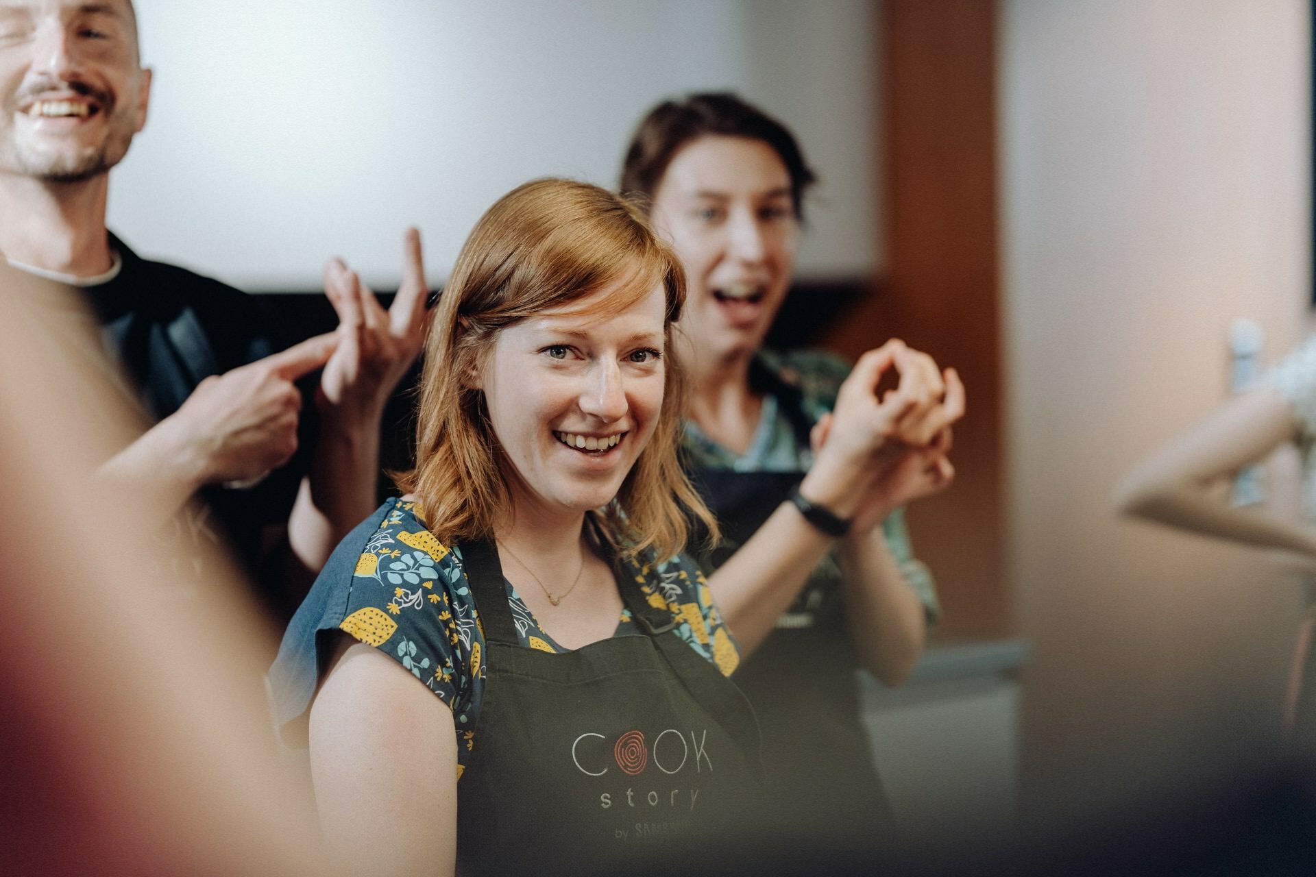 A woman with shoulder-length reddish-brown hair smiles warmly, wearing a black apron printed with "COOK History." Two people in the background, also dressed in aprons, make animated gestures and appear to be immersed in a lively conversation, perfect for a photo essay of the events. 