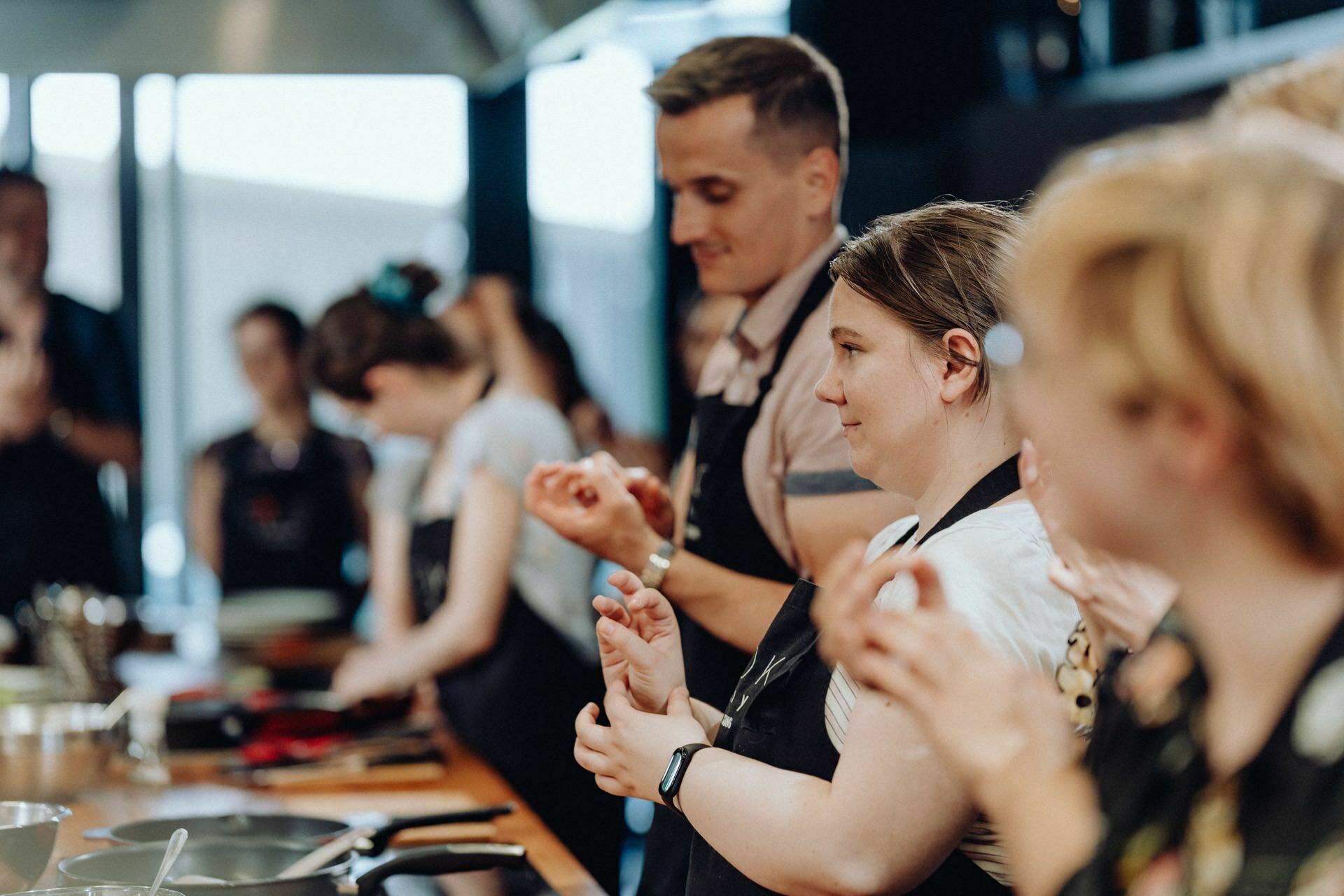 A group of people dressed in aprons stand around the kitchen counter and get busy cooking. Some are clapping, while others are focused on preparing food. The background is blurred, highlighting the participants and their activities, which this event photography captures perfectly.  