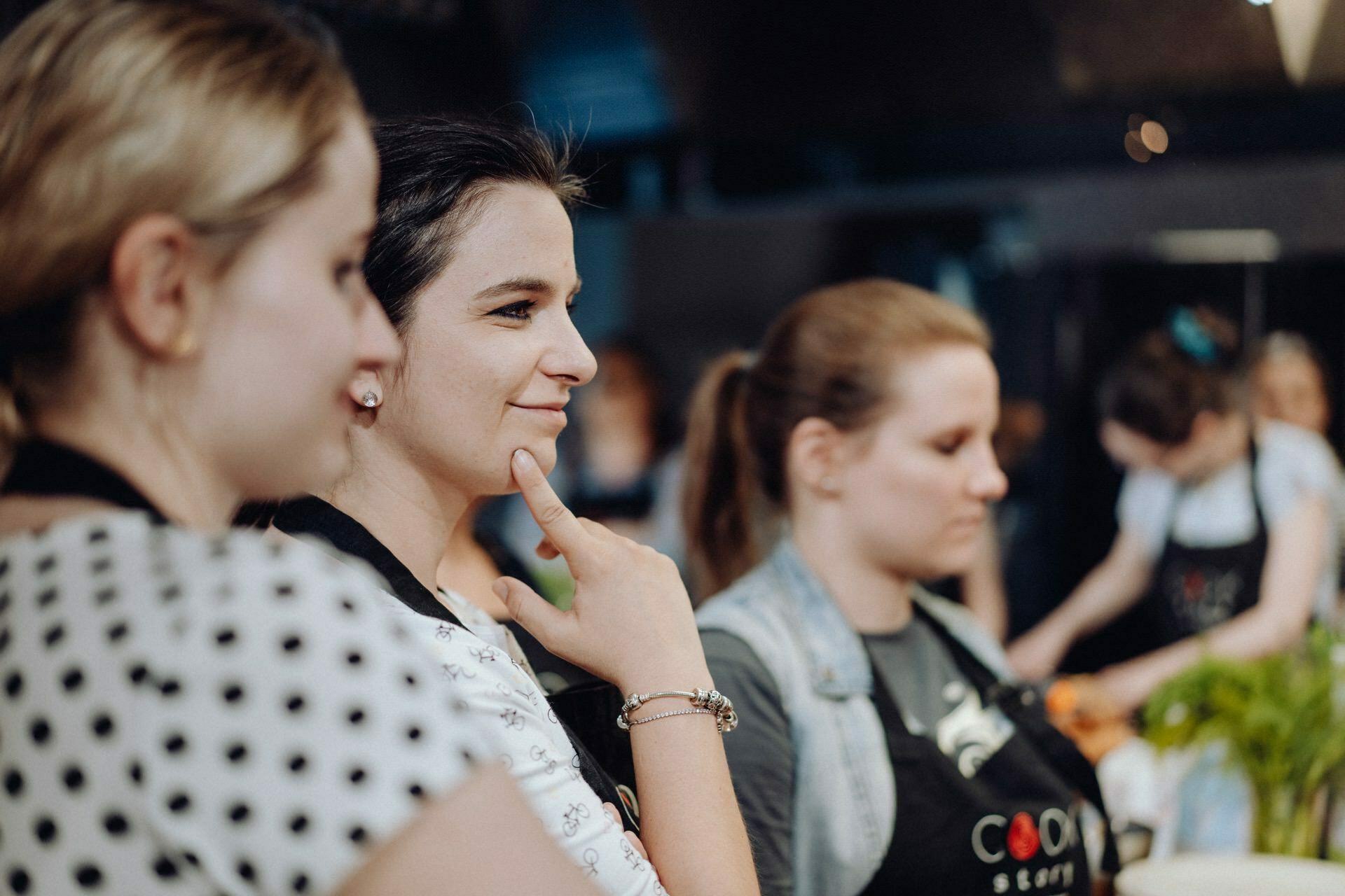 Three women wearing aprons during a cooking class. The woman in the foreground is thoughtfully touching her chin, the middle woman is smiling, and the third woman seems focused on the task at hand. In the background is a blur of kitchen activity - the perfect event photo captured by a talented event photographer warsaw.  