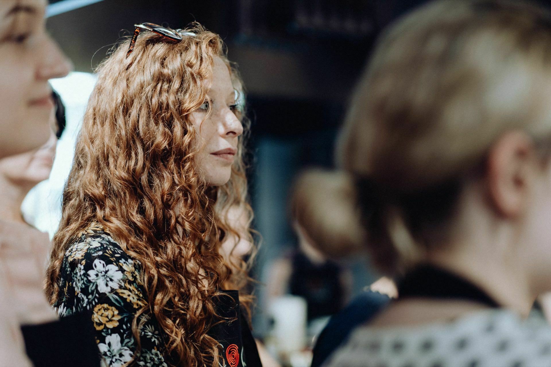 A woman with long curly red hair and sunglasses on her head listens intently during what appears to be a group event or workshop depicted in this event photo. She is wearing a floral patterned top and is surrounded by other people whose faces are blurred and out of focus. 