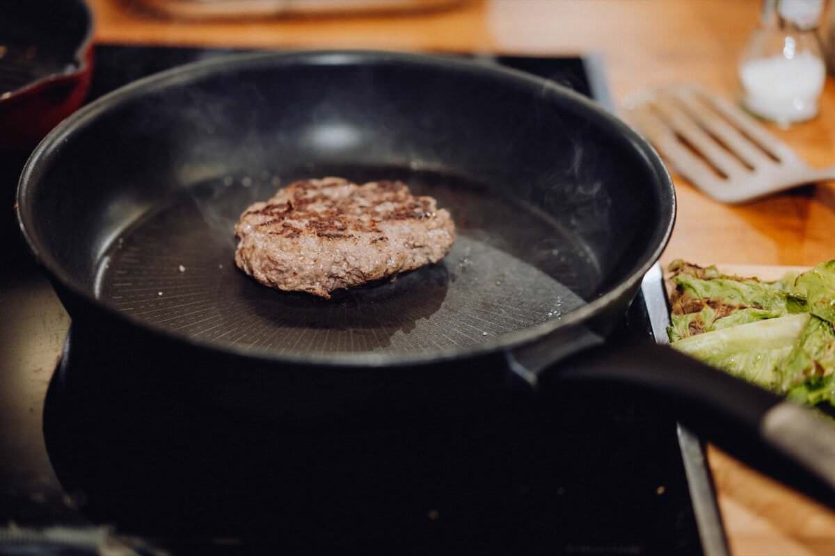 Kotlet hamburgerowy smaży się na czarnej patelni na kuchence. W tle leży szpatułka, a po prawej stronie patelni znajdują się zielone warzywa liściaste. To tak, jakby fotograf eventowy warszawa uchwycił ten spokojny kuchenny moment podczas sesji fotografia eventowa.