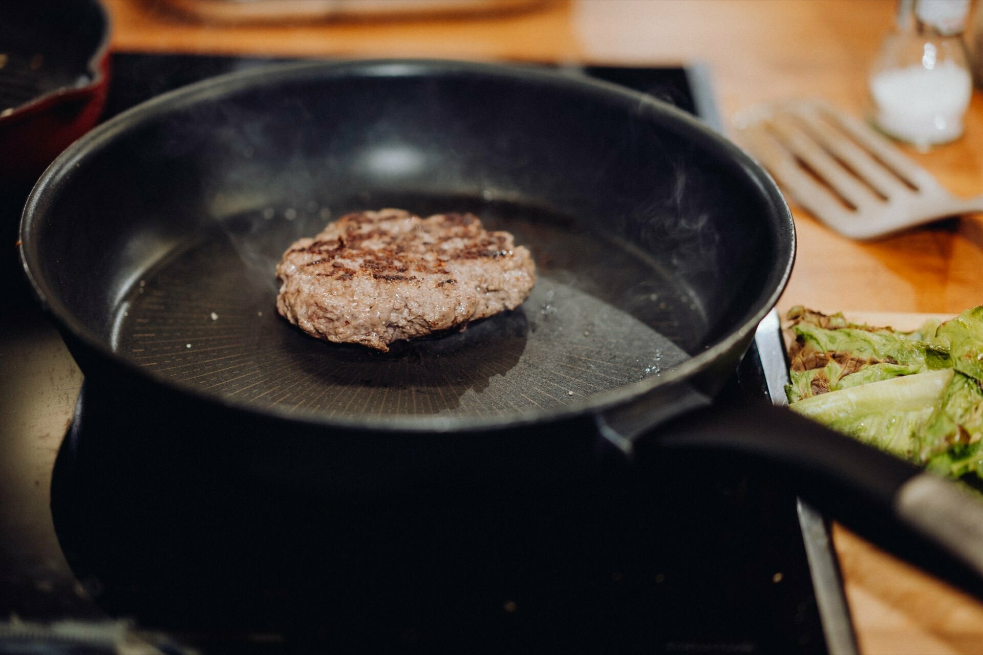 A hamburger cutlet is frying in a black pan on the stove. A spatula lies in the background, and to the right of the pan are green leafy vegetables. It's as if an event photographer warsaw captured this peaceful kitchen moment during an event photography session.  