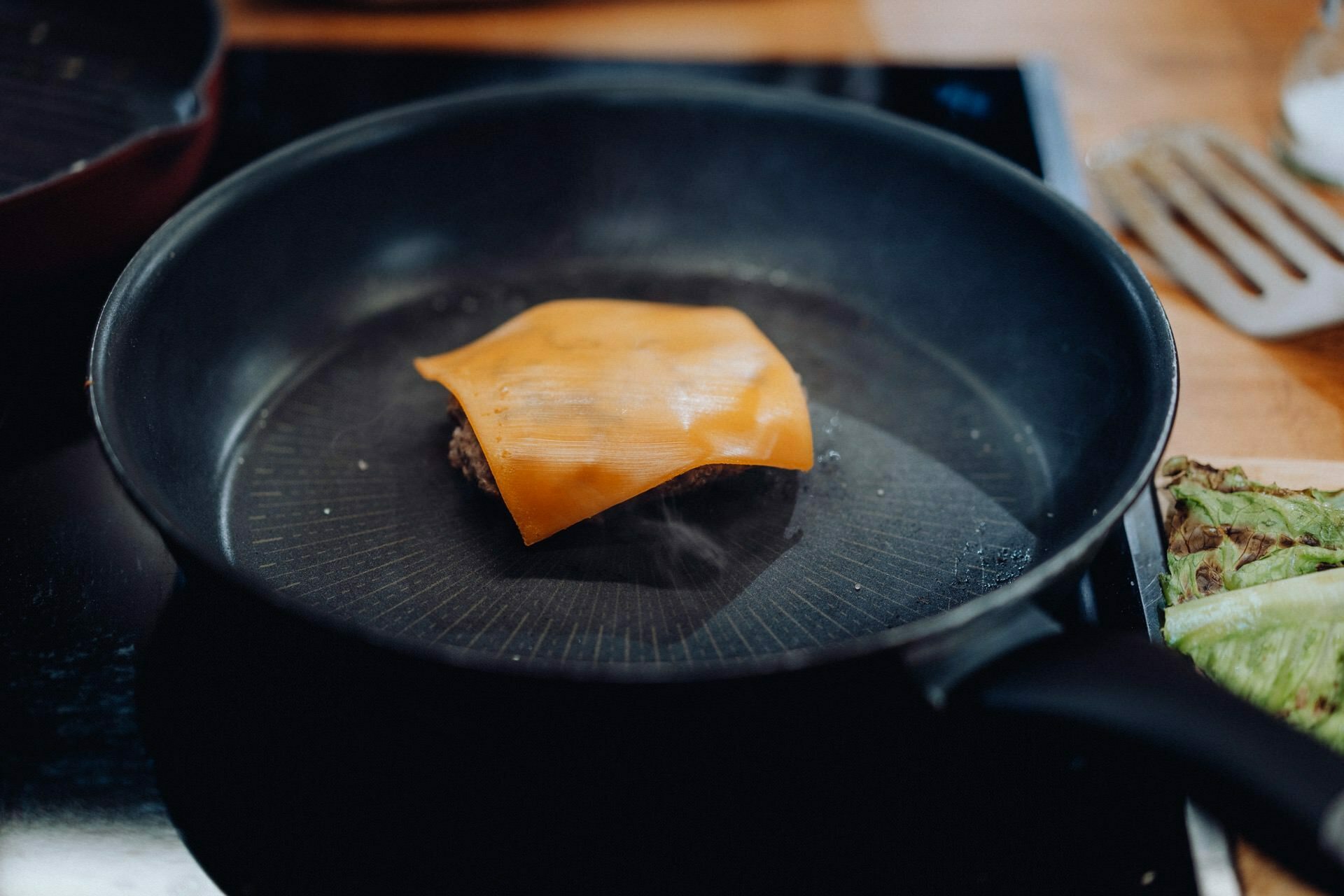 A slice of cheddar cheese melting into a burger patty on a black pan set on a cooktop, captured with the precision of an event photographer warsaw. In the background, lettuce and a metal spatula can be seen on a wooden countertop. 